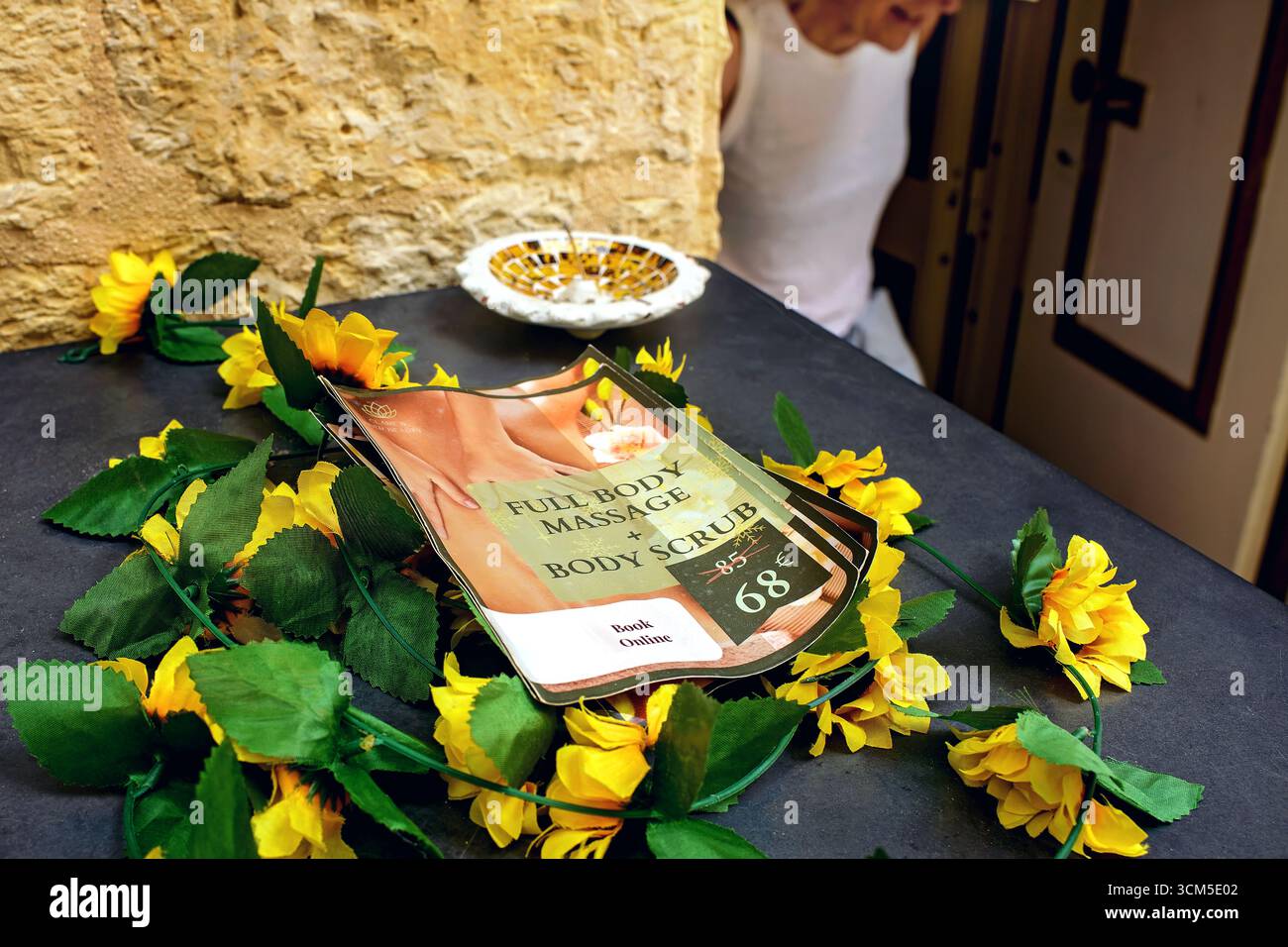 La Valletta, Malta - 08.26.2025: Primo piano di un tavolo con volantino promozionale, fiori gialli e silhouette del visitatore vicino all'ingresso del salone di massaggi. Foto Stock