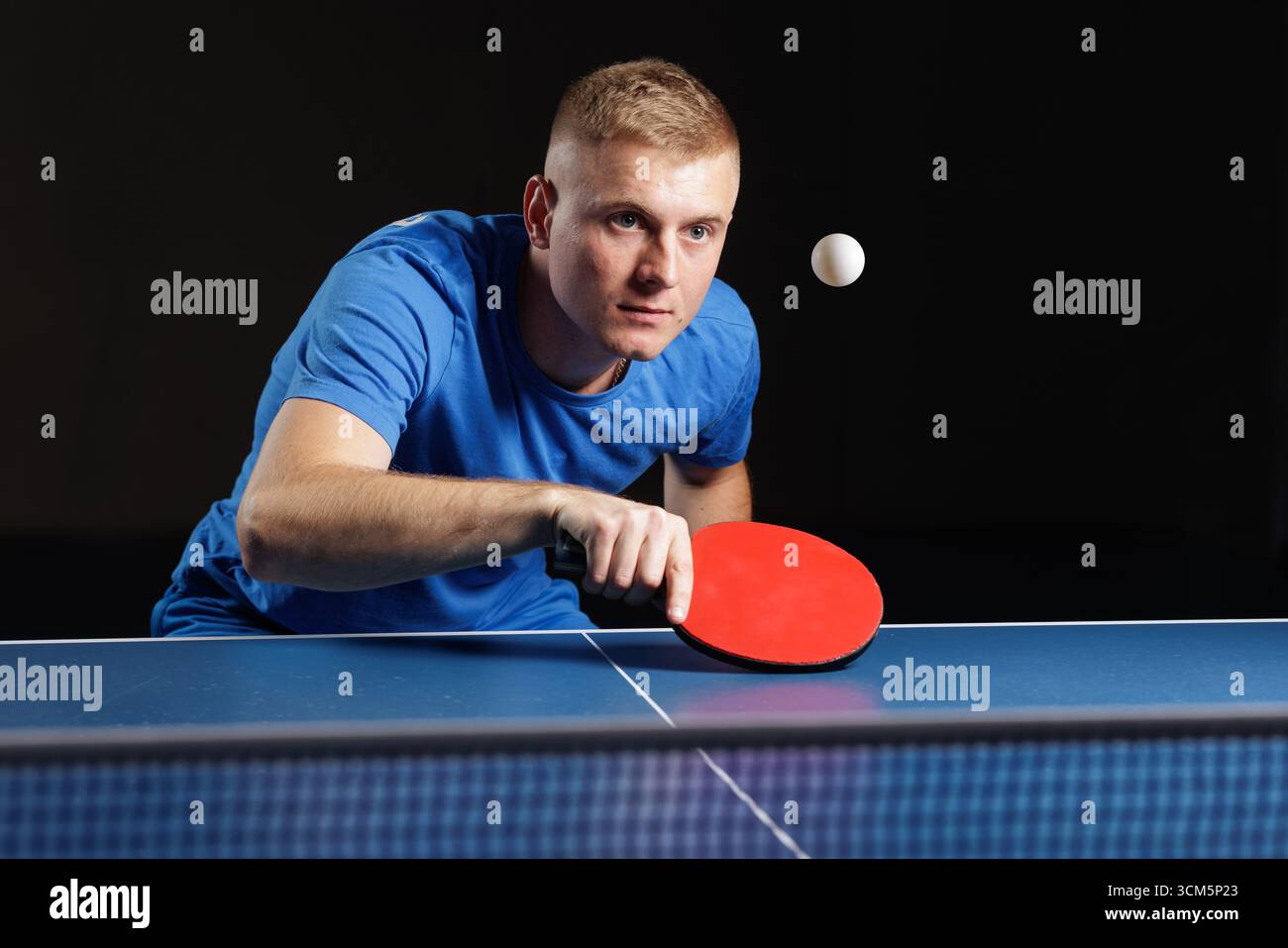 Un atleta maschile specializzato si prepara a servire su un campo da ping-pong al coperto con pagaia rossa. Perfetto per concetti di formazione, coaching e concorrenza. Foto Stock