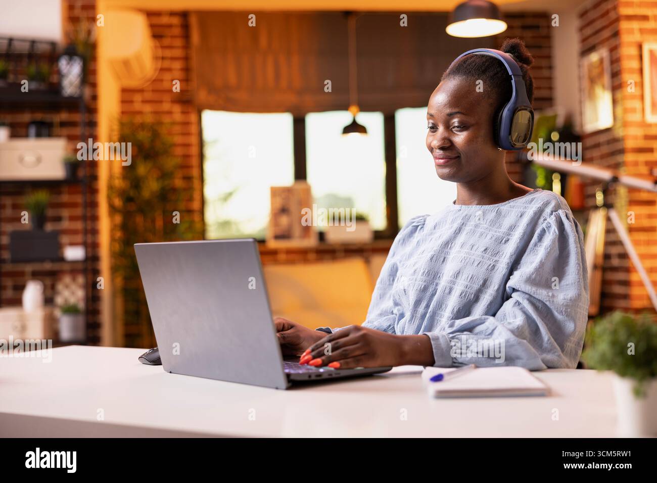 La freelance femminile nera ascolta la musica con le cuffie mentre digita sul dispositivo digitale. Donna afroamericana felice che indossa un auricolare wireless siede alla scrivania, studia e prepara la presentazione sul laptop. Foto Stock