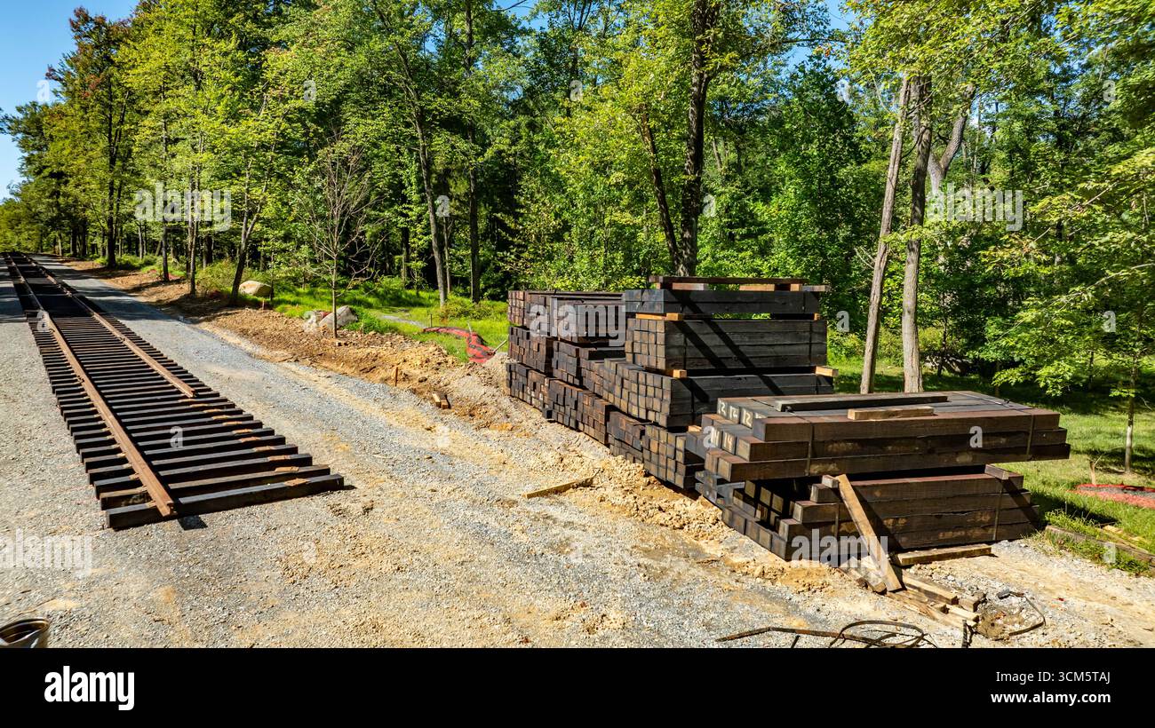Le fascette ferroviarie in legno sono impilate ordinatamente accanto a un sentiero di ghiaia che diventerà parte della nuova ferrovia. Gli alberi circostanti offrono uno sfondo verde lussureggiante Foto Stock
