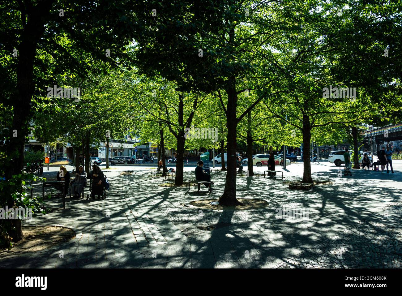 Le persone si rilassano all'ombra dei vibranti alberi verdi a Kottbusser Tor, Berlino. La luce solare soffusa crea un bellissimo motivo a contrasto elevato sul terreno, Foto Stock