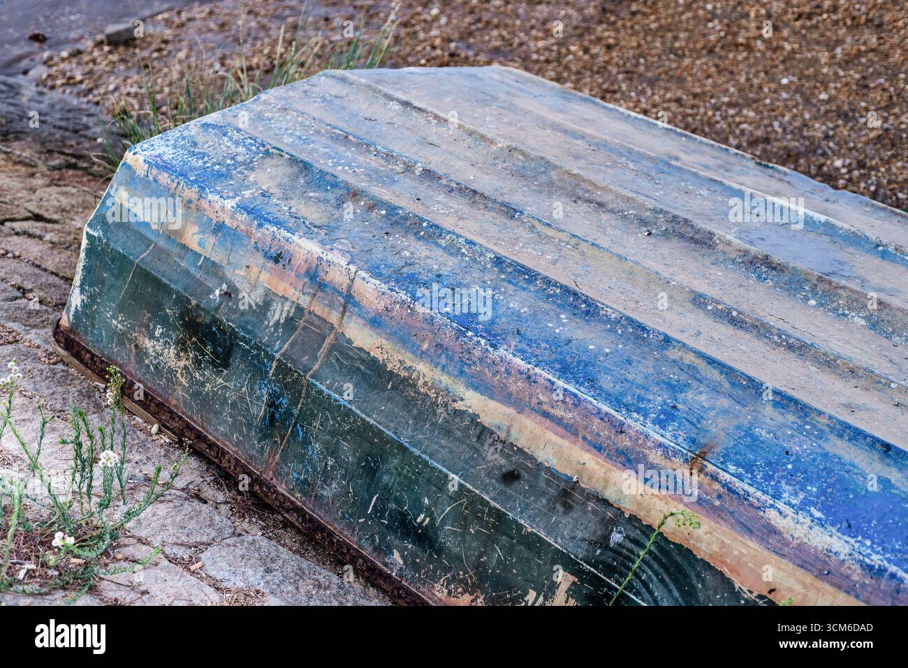 Barco abandonado boca abajo en la orilla del Río Tinto, con pintura desgastada y óxido visible, rodeado de agua tranquila y sobre suelo de granito. Foto Stock