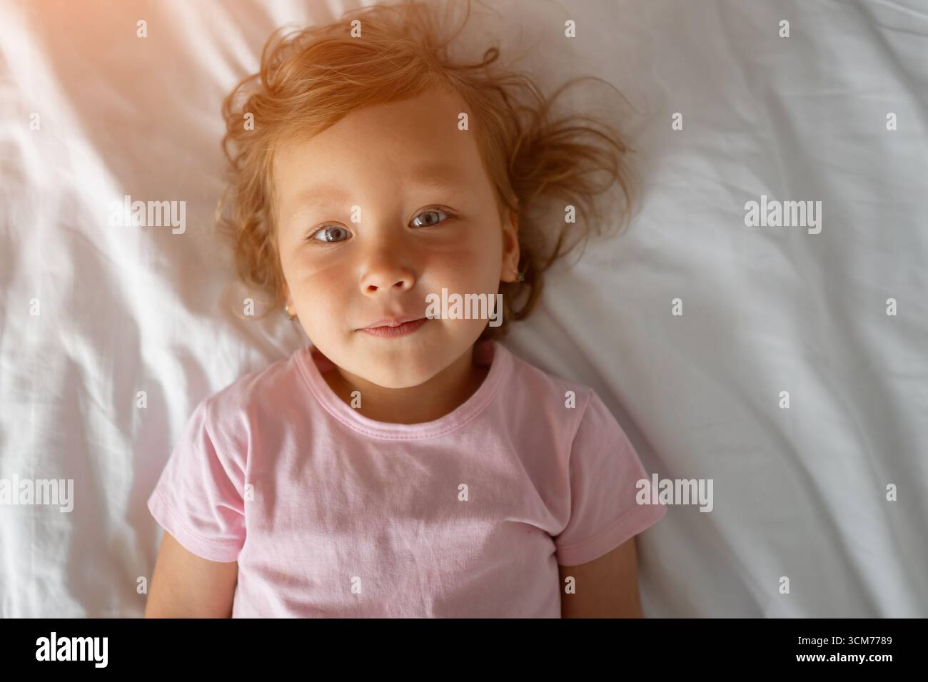Tenera bambina che giace su biancheria da letto bianca e indossa una t shirt rosa Foto Stock