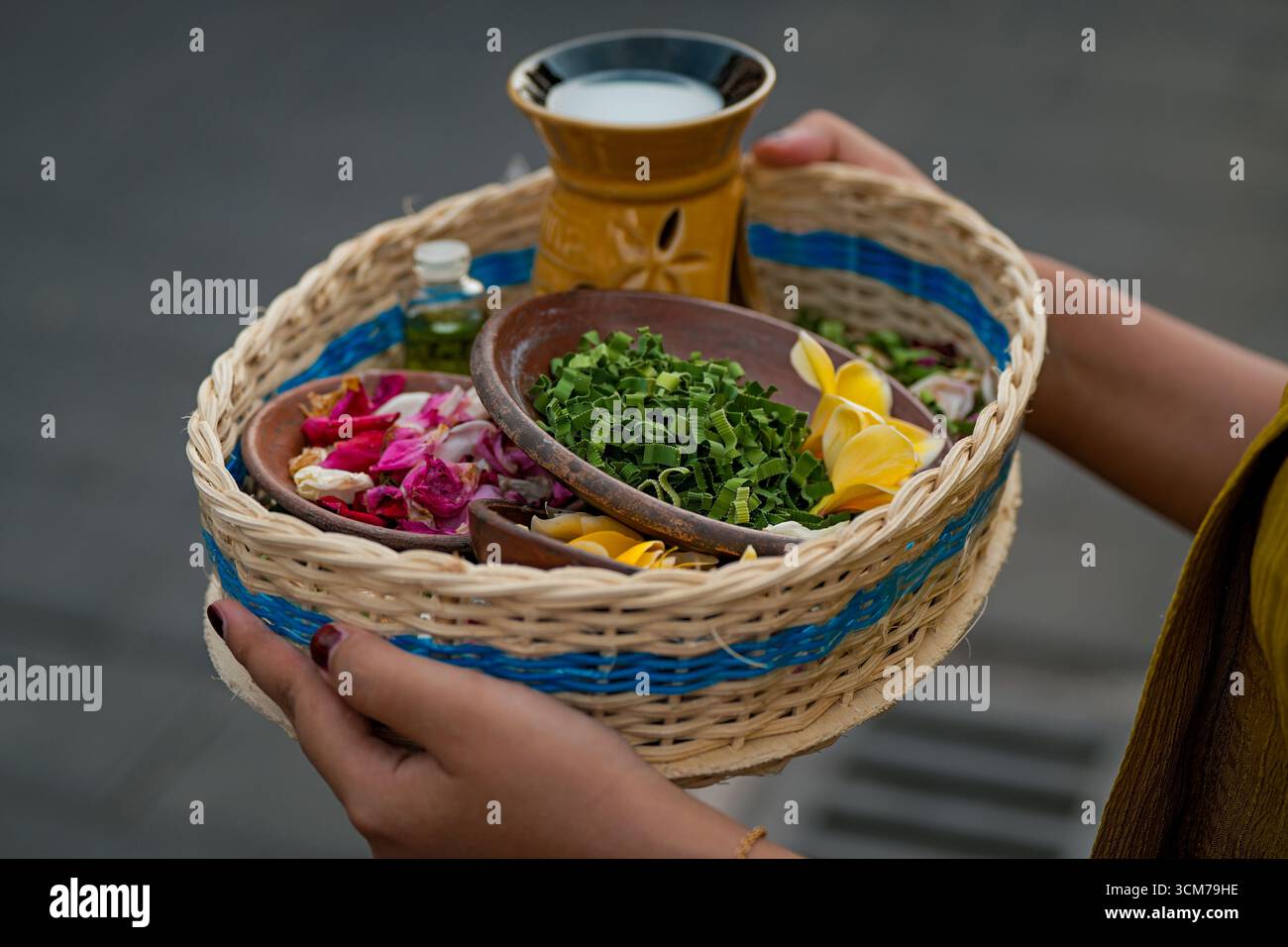 Un cestino in tessuto pieno di fiori colorati, erbe aromatiche e una piccola bottiglia, tenuta da una persona, con una tazza decorativa sullo sfondo Foto Stock