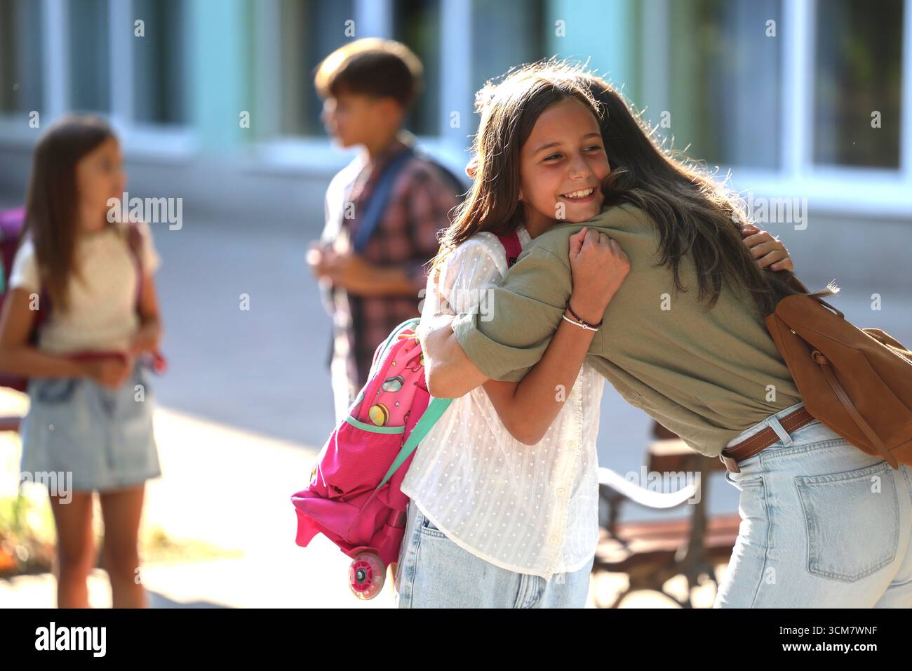 La gioiosa riunione ha luogo quando la madre abbraccia sua figlia dopo la scuola. Altri bambini giocano e socializzano in sottofondo nelle giornate di sole Foto Stock