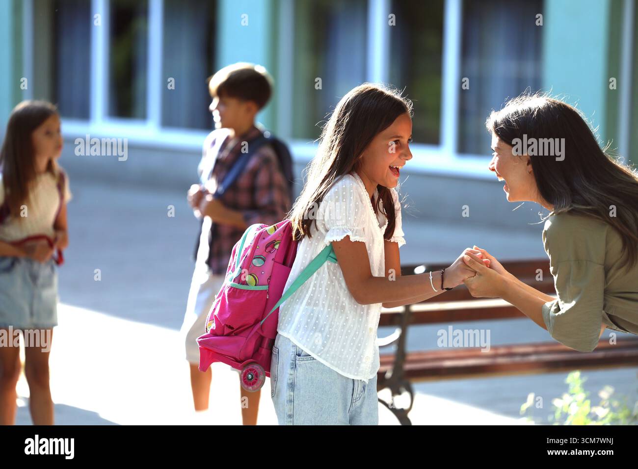 Madre eccitata ha interagito con sua figlia felice, godendosi il loro tempo insieme fuori scuola mentre altri bambini camminano in background Foto Stock