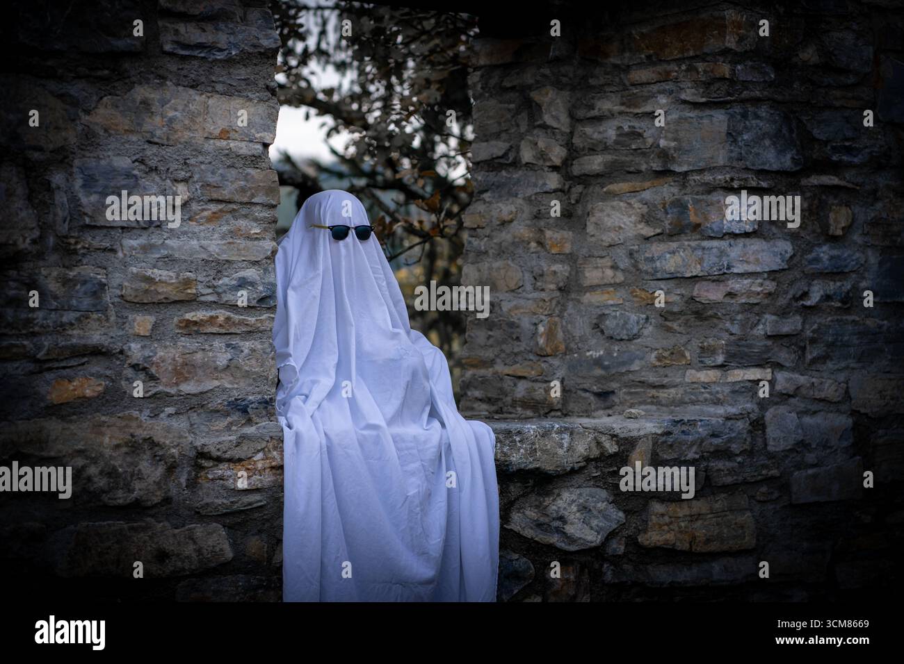 Il personaggio, avvolto in un foglio bianco che scorre, si trova su una parete di pietra con eleganti occhiali da sole, creando un'intrigante combinazione di aspetto spettrale Foto Stock