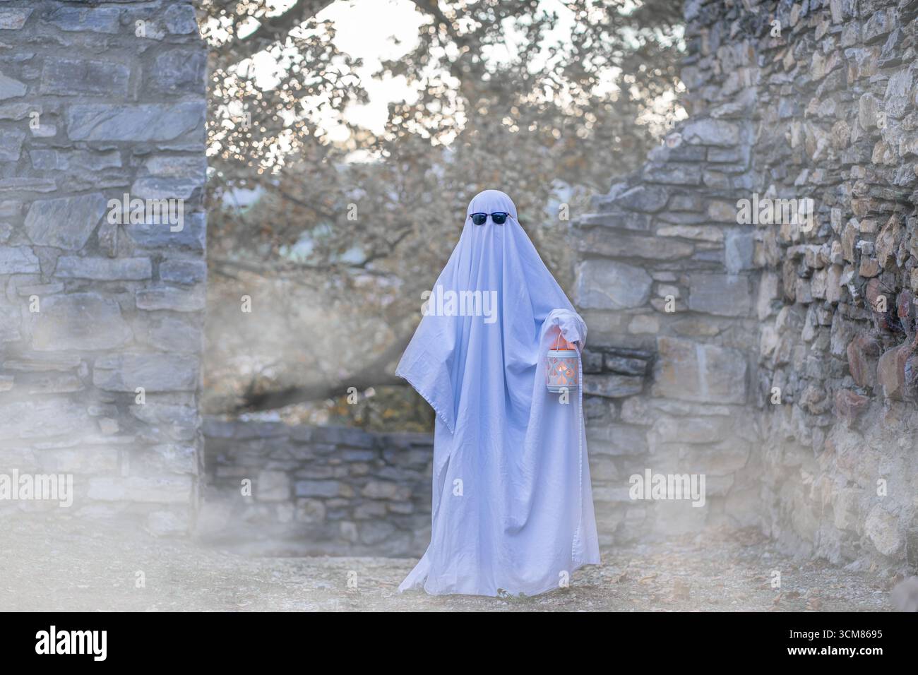 Un bambino in un foglio bianco che scorre cammina lungo una parete di pietra indossando occhiali da sole e tenendo in mano una lanterna luminosa nel mezzo del fumo bianco Foto Stock