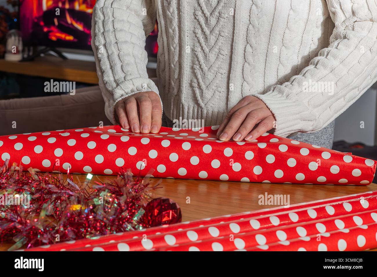 Donna che avvolge i regali di Natale in carta a pois rossa vicino all'albero decorato. Preparazione per le vacanze, tempo in famiglia e atmosfera festosa a casa Foto Stock