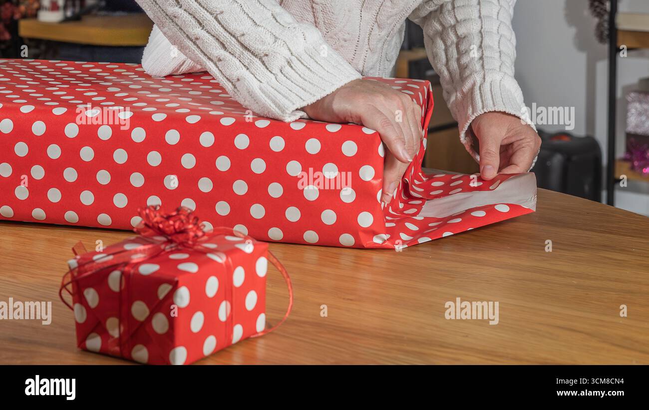 Donna che avvolge i regali di Natale in carta a pois rossa vicino all'albero decorato. Preparazione per le vacanze, tempo in famiglia e atmosfera festosa a casa Foto Stock