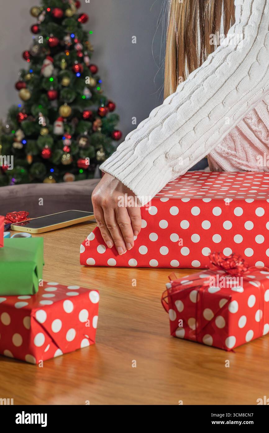 Donna che avvolge i regali di Natale in carta a pois rossa vicino all'albero decorato. Preparazione per le vacanze, tempo in famiglia e atmosfera festosa a casa Foto Stock