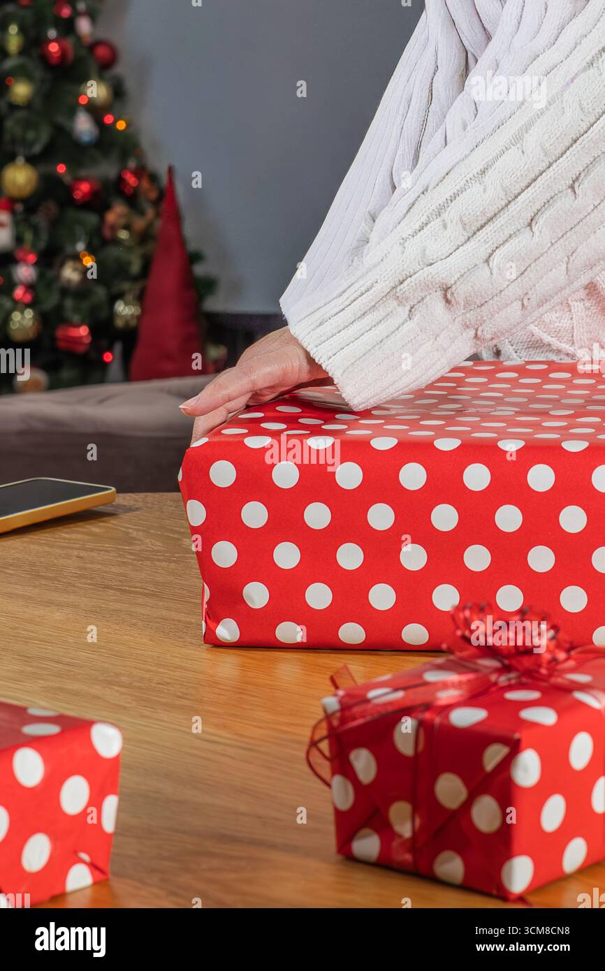Donna che avvolge i regali di Natale in carta a pois rossa vicino all'albero decorato. Preparazione per le vacanze, tempo in famiglia e atmosfera festosa a casa Foto Stock