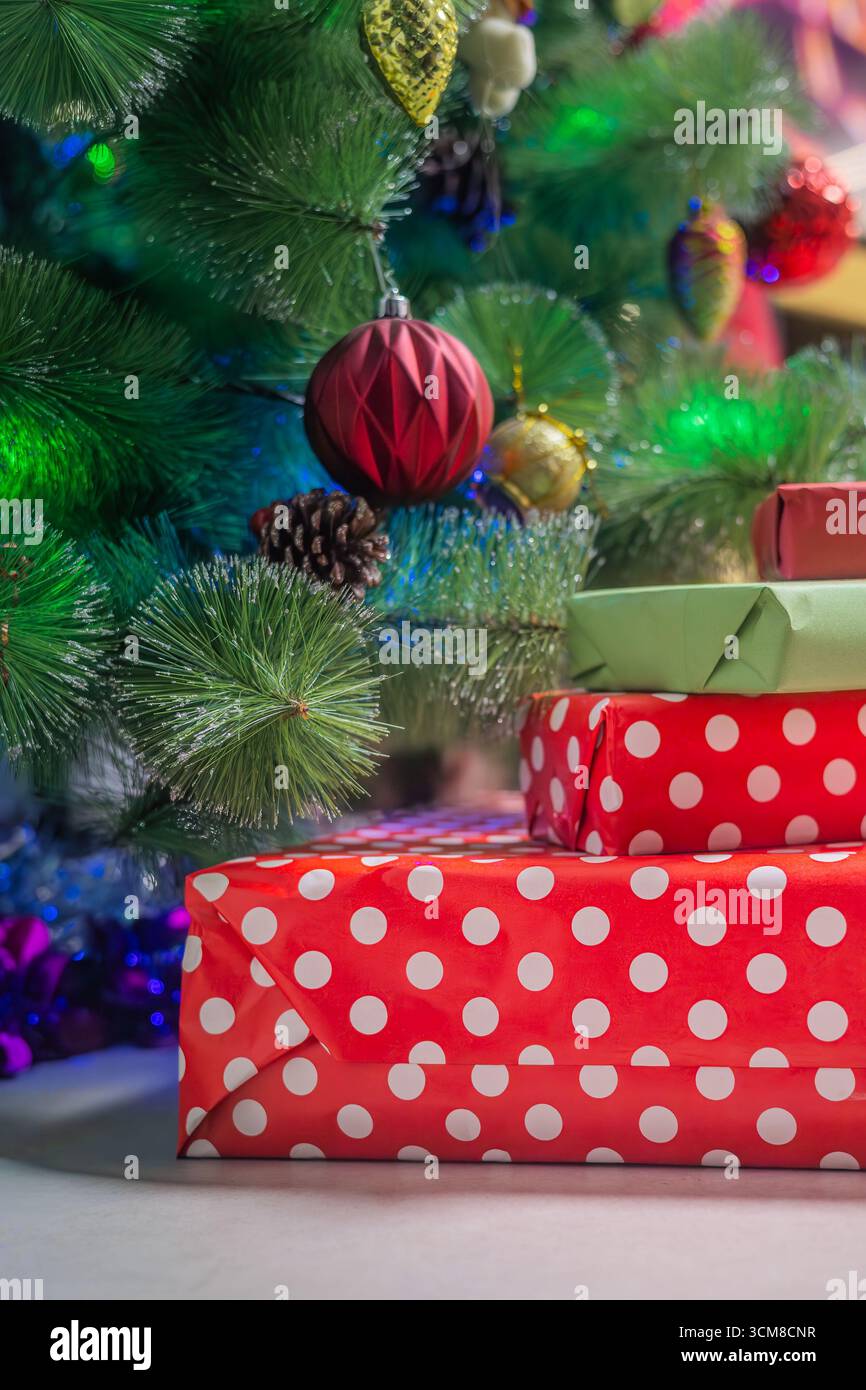 Pila di regali di Natale avvolti in rosso e verde sotto l'albero decorato con luci festive. Festeggiamenti, famiglia, atmosfera accogliente Foto Stock