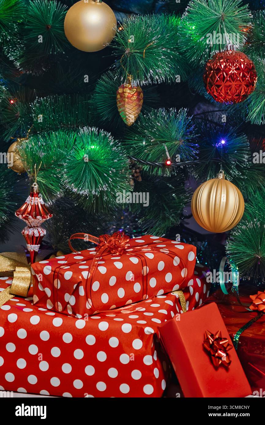 Pila di regali di Natale avvolti in rosso e verde sotto l'albero decorato con luci festive. Festeggiamenti, famiglia, atmosfera accogliente Foto Stock