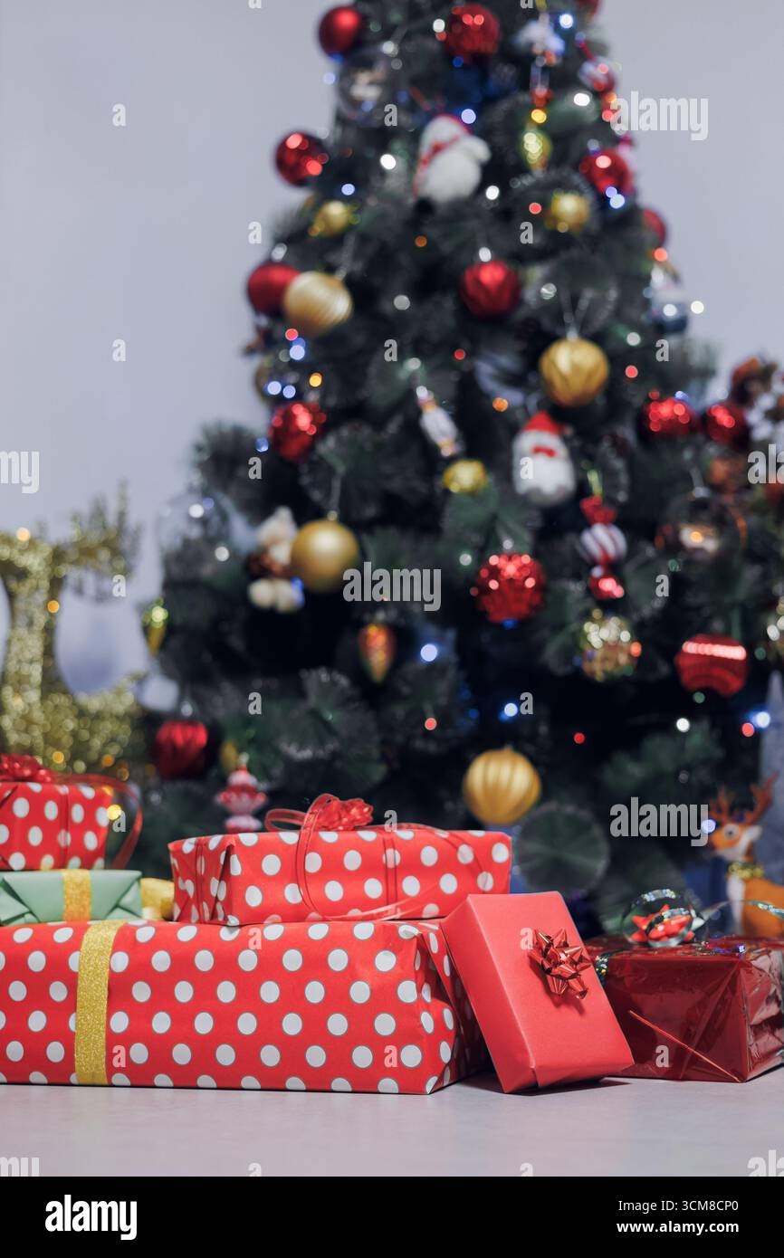 Pila di regali di Natale avvolti in rosso e verde sotto l'albero decorato con luci festive. Festeggiamenti, famiglia, atmosfera accogliente Foto Stock