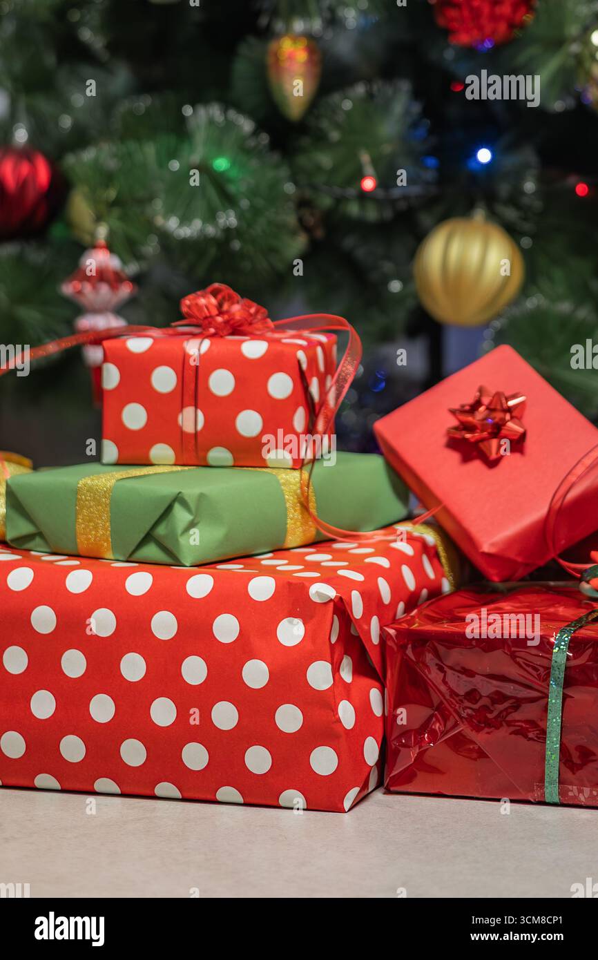 Pila di regali di Natale avvolti in rosso e verde sotto l'albero decorato con luci festive. Festeggiamenti, famiglia, atmosfera accogliente Foto Stock