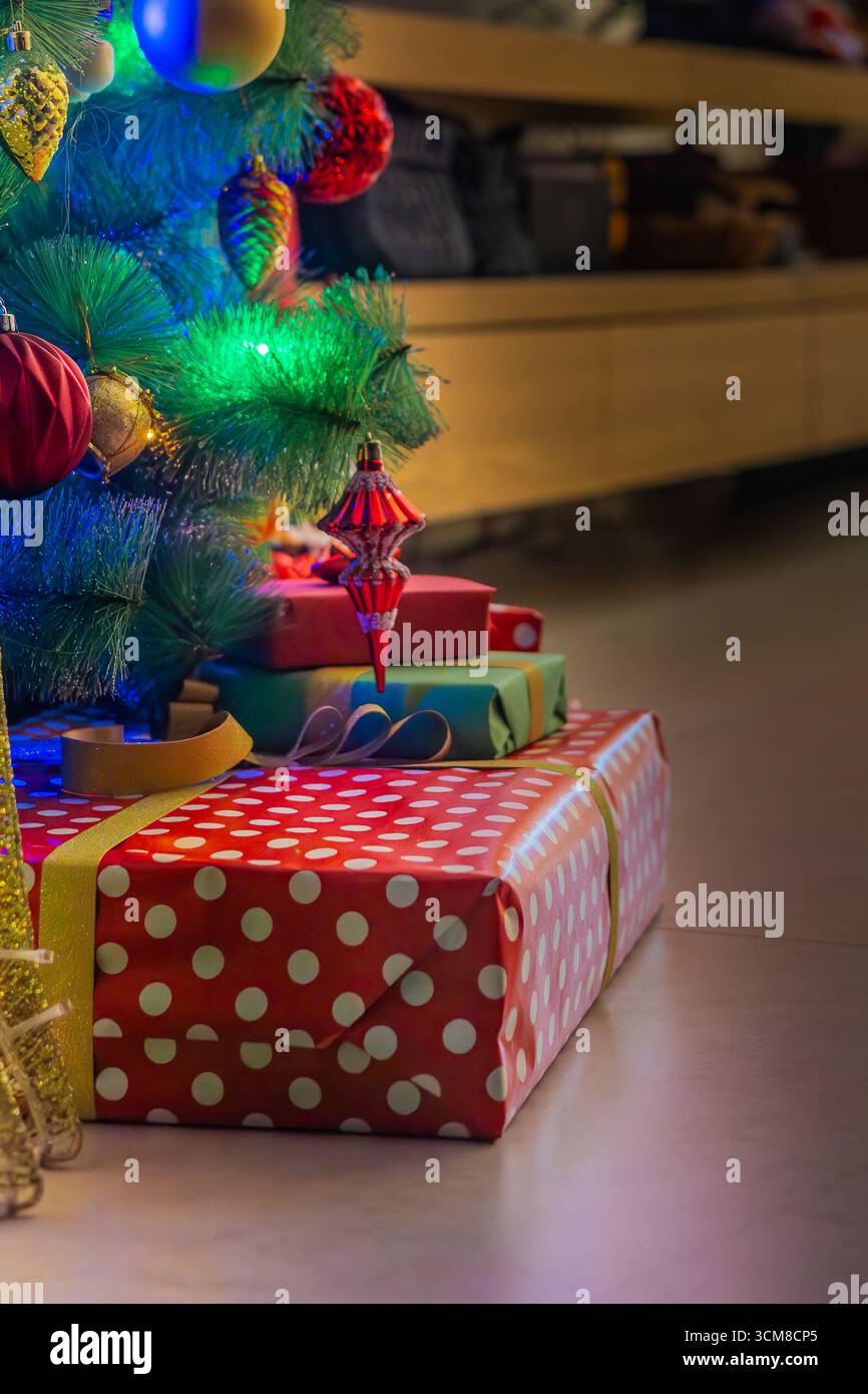 Pila di regali di Natale avvolti in rosso e verde sotto l'albero decorato con luci festive. Festeggiamenti, famiglia, atmosfera accogliente Foto Stock