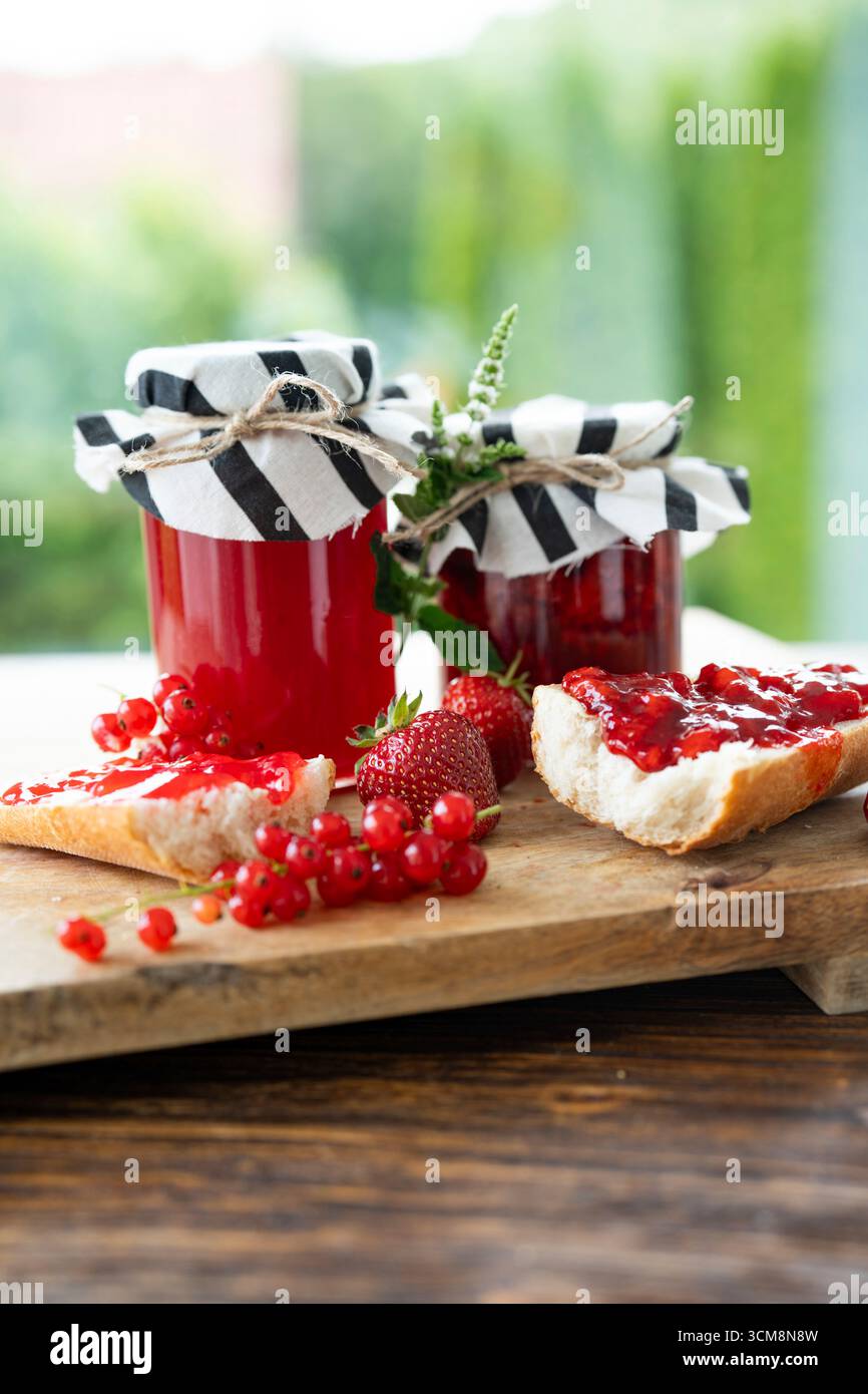 Confettura fatta in casa di fragole e ribes in vaso. Bacche rosse e baguette croccanti con frutta fresca spalmata su un tagliere di legno. Primo piano verticale con spazio per il testo. Foto Stock