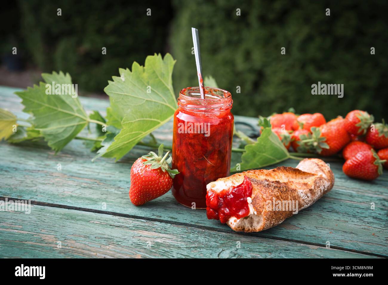 Marmellata di fragole fatta in casa in un barattolo con baguette fresca su un vecchio tavolo di legno all'aperto. Spalmabile a base di frutta fresca di stagione. Primo piano. Foto Stock