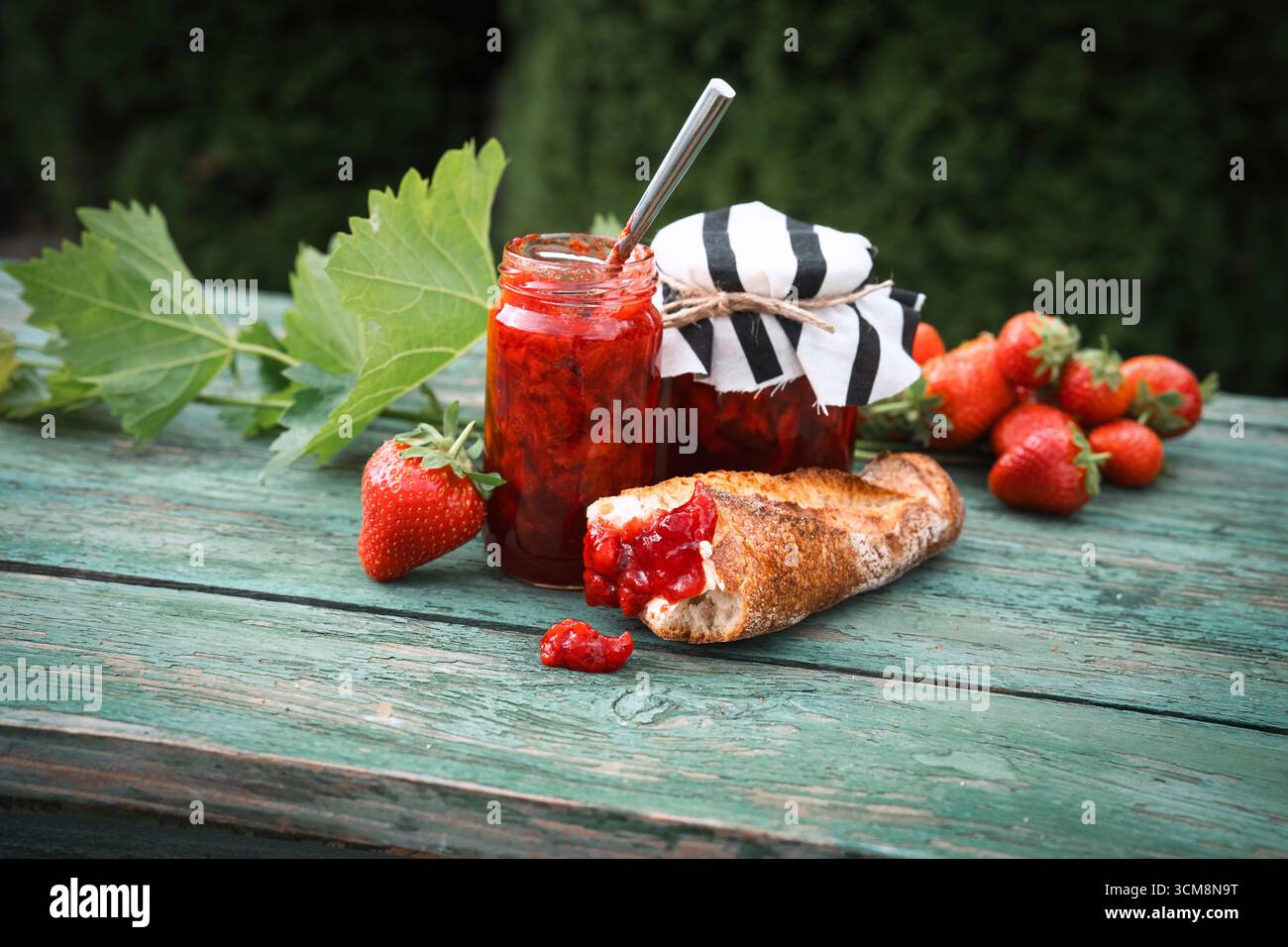 Marmellata di fragole fatta in casa in un barattolo con baguette fresca su un vecchio tavolo di legno all'aperto. Spalmabile a base di frutta fresca di stagione. Primo piano. Foto Stock