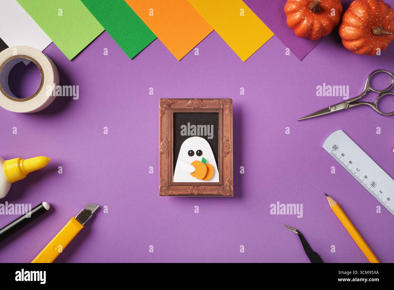 La carta di Halloween ritrae un ritratto di un fantasma che tiene in mano una zucca. Carta colorata, forbici, colla, righello, nastro, coltello e zucche sul backgroun viola Foto Stock