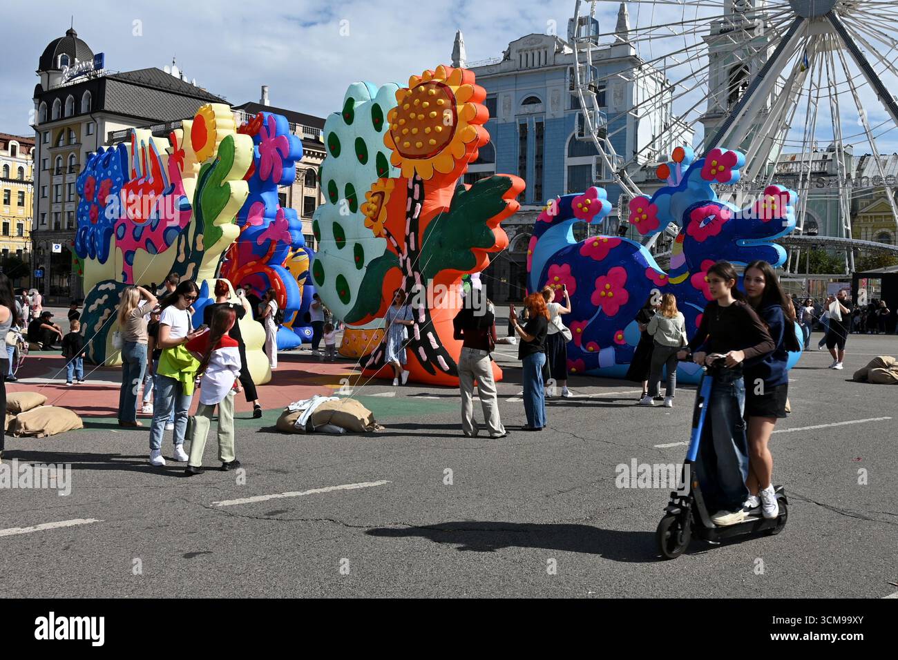 Kiev, Ucraina. 13 settembre 2025. I residenti di Kiev e gli ospiti della capitale camminano lungo Piazza Kontraktova sullo sfondo della ruota panoramica e l'installazione di sculture gonfiabili del duo artistico newyorkese "Chiaozza" (Adam Frezza e Terri Chiaode) ispirate al lavoro dell'artista Ucraina Maria Prymachenko, nel mezzo dell'invasione russa dell'Ucraina. L'Ucraina continua a difendersi dall'aggressione militare russa per il quarto anno dopo l'invasione del 24 febbraio 2022, con bombardamenti del territorio ucraino che continuano quotidianamente. Credito: SOPA Images Limited/Alamy Live News Foto Stock
