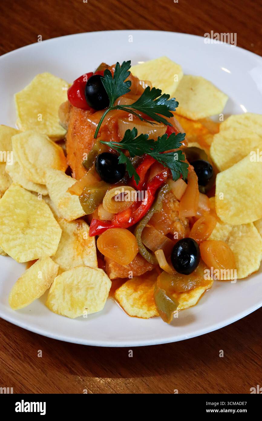 il tradizionale merluzzo, il pezzo principale del pesce è coperto da un colorato mix di cipolle saltate e peperoni, patatine fritte, guarnite con olive nere intere Foto Stock