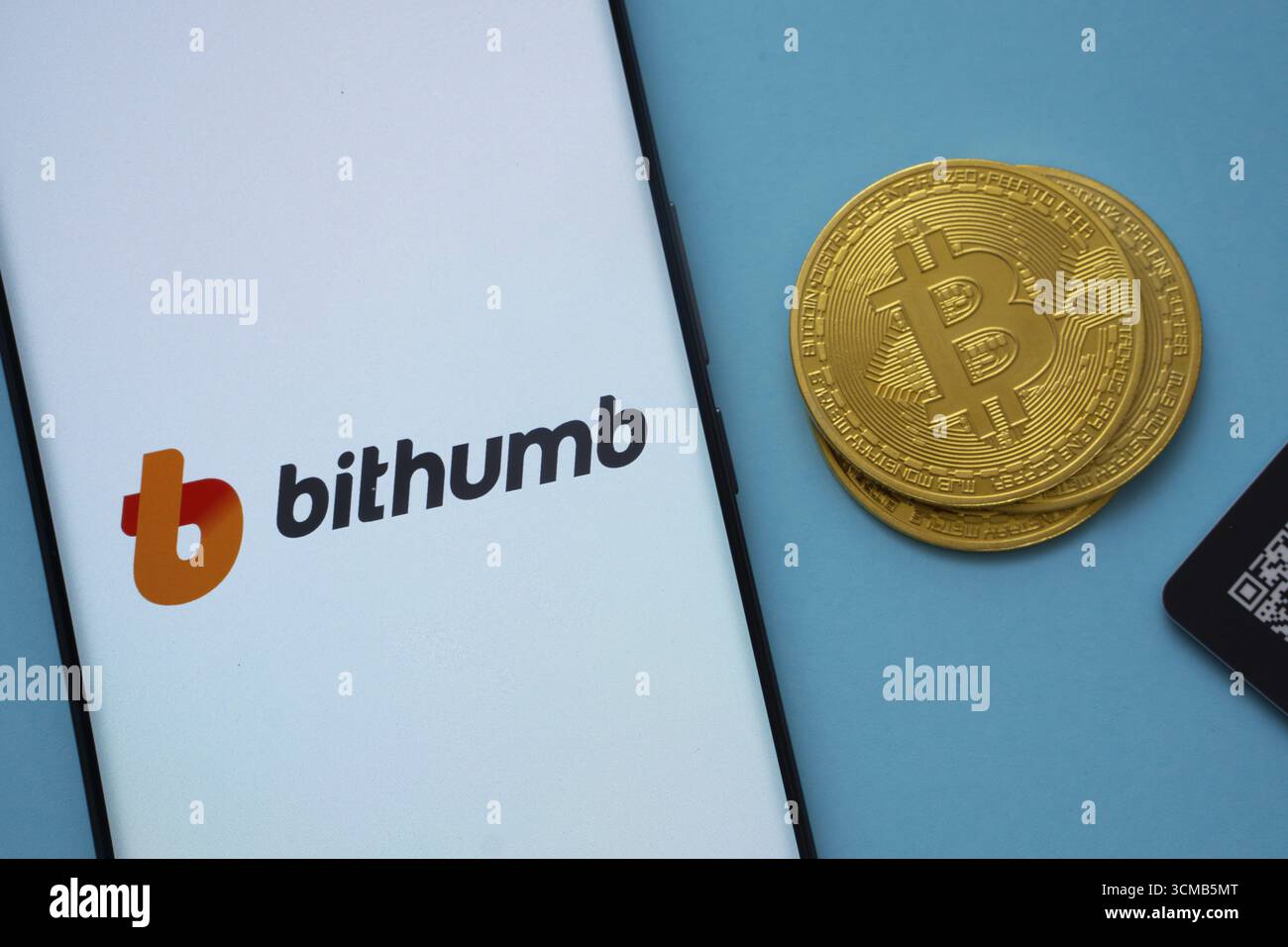 Dhaka, Bangladesh - 7 maggio 2025: Logo Bithumb sullo schermo dello smartphone vicino alla moneta Bitcoin e codice QR su sfondo blu Foto Stock