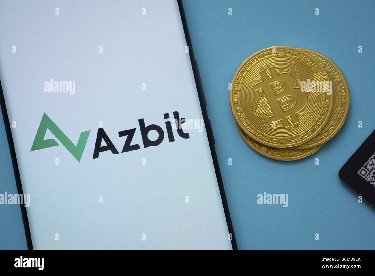 Dacca, Bangladesh - 2 giugno 2025: Logo Azbit sullo schermo dello smartphone vicino alla moneta Bitcoin e codice QR su sfondo blu Foto Stock