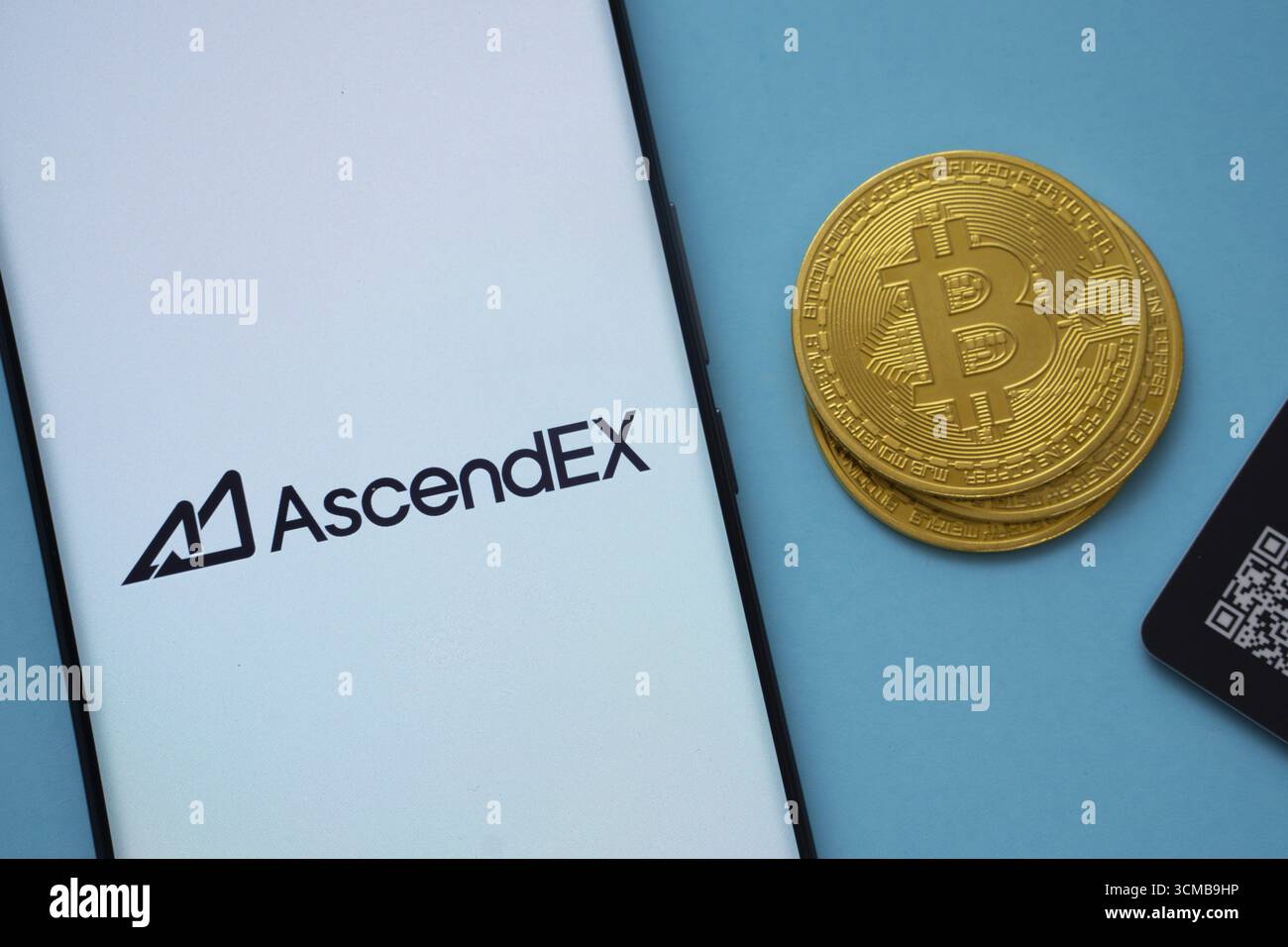Dhaka, Bangladesh - 2 giugno 2025: Logo AscendEX sullo schermo dello smartphone vicino a Bitcoin Coin e codice QR su sfondo blu Foto Stock