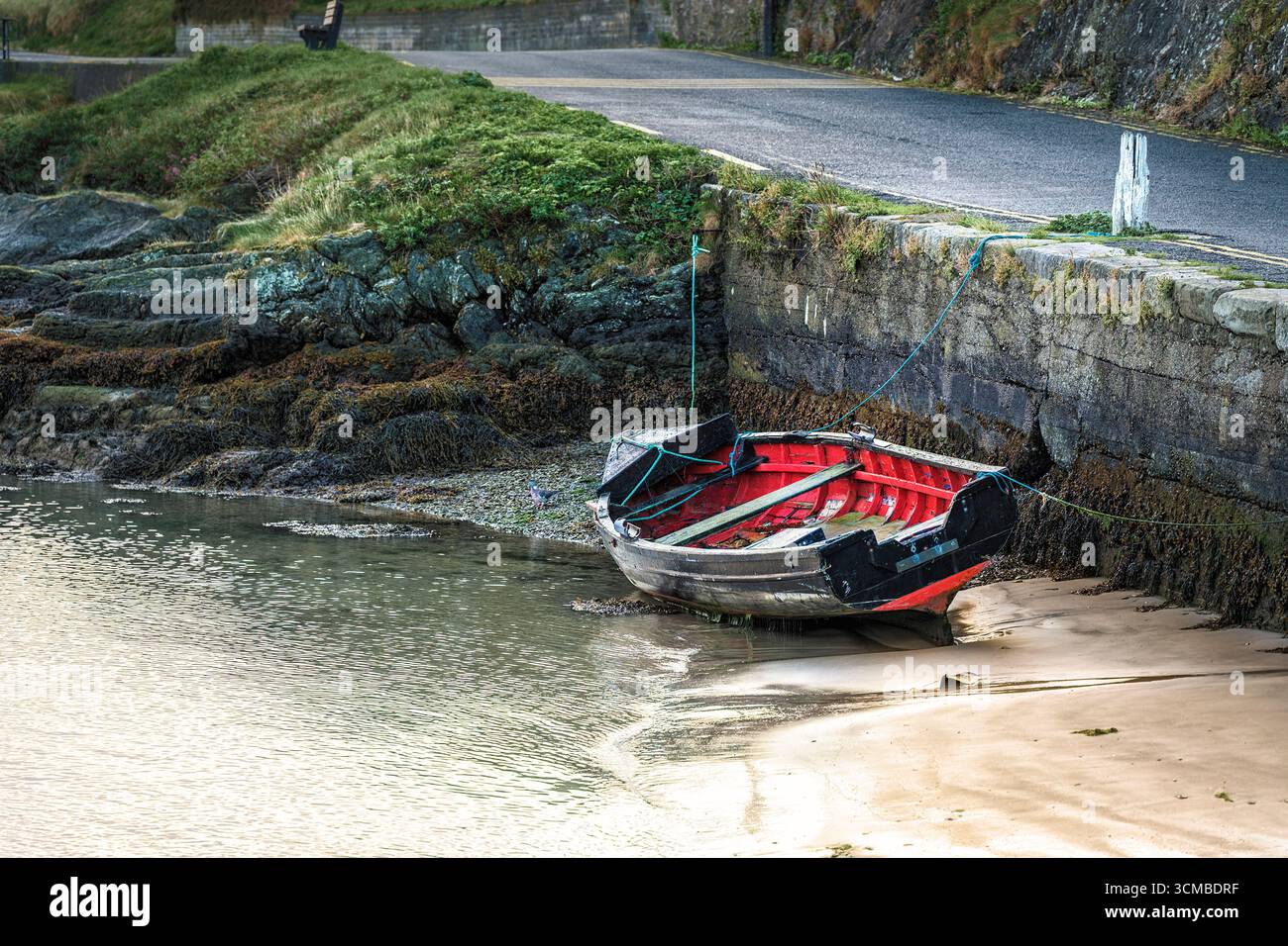 La barca a remi si appoggia sulla sabbia di Wicklow Harbour con la bassa marea, incorniciata da una costa rocciosa, una riva erbosa e un molo di pietra. Foto Stock