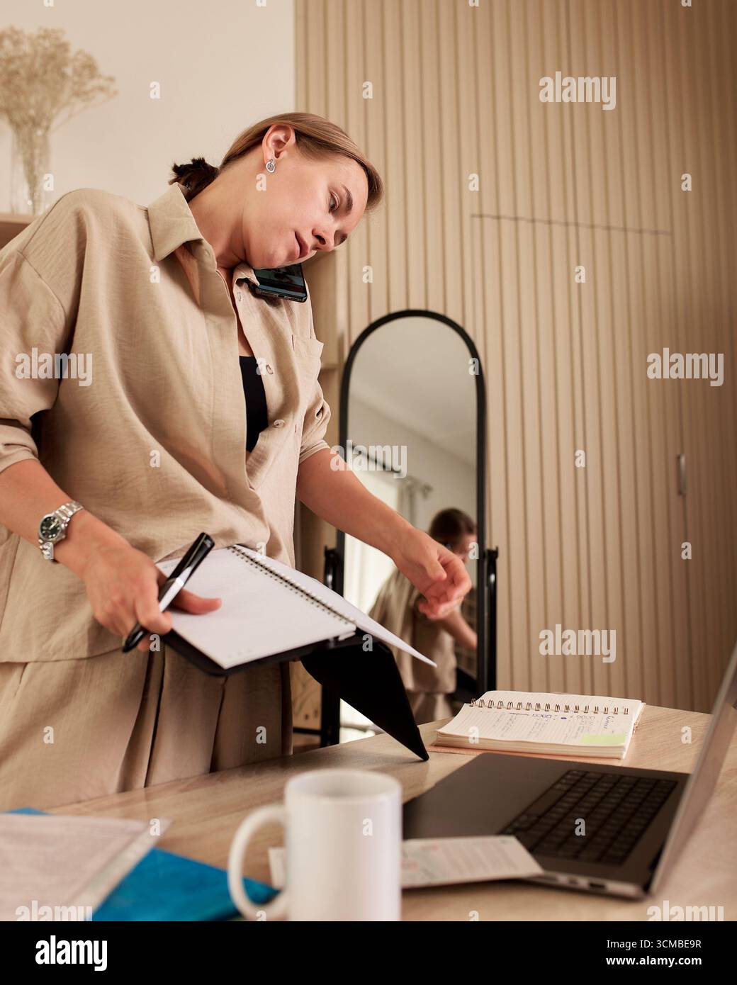 Un adulto professionista che lavora in multitasking in un ufficio domestico, impegnato in telefonate e documenti. Lo spazio di lavoro è dotato di un laptop, di notebook aperti e di un cof Foto Stock