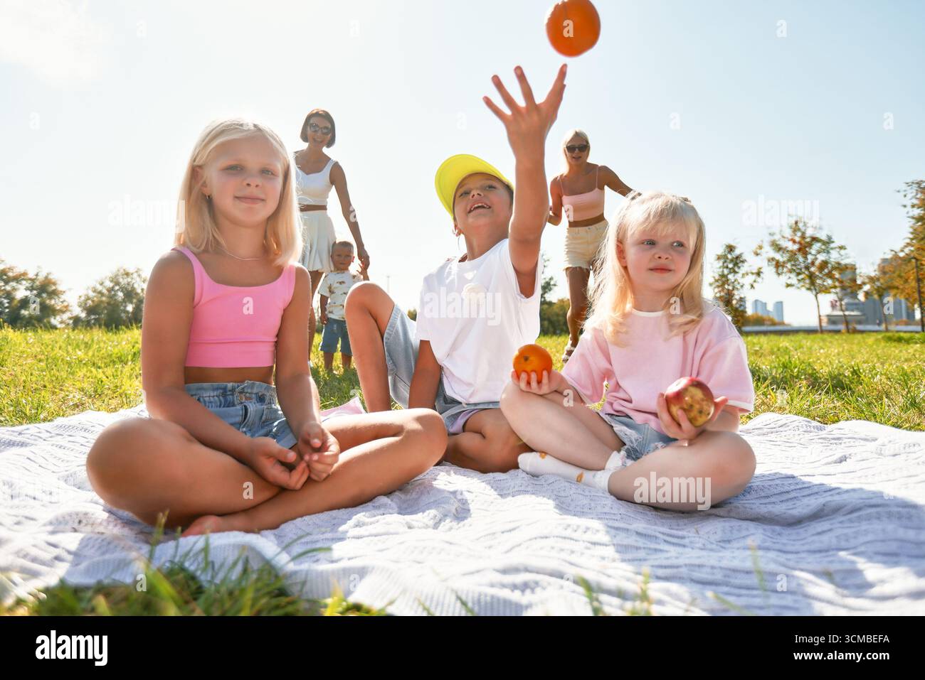 Una divertente riunione di famiglia in un parco pittoresco in una giornata di sole. I bambini si siedono su una coperta da picnic, godendosi il momento e giocando con pezzi di frutta, W Foto Stock