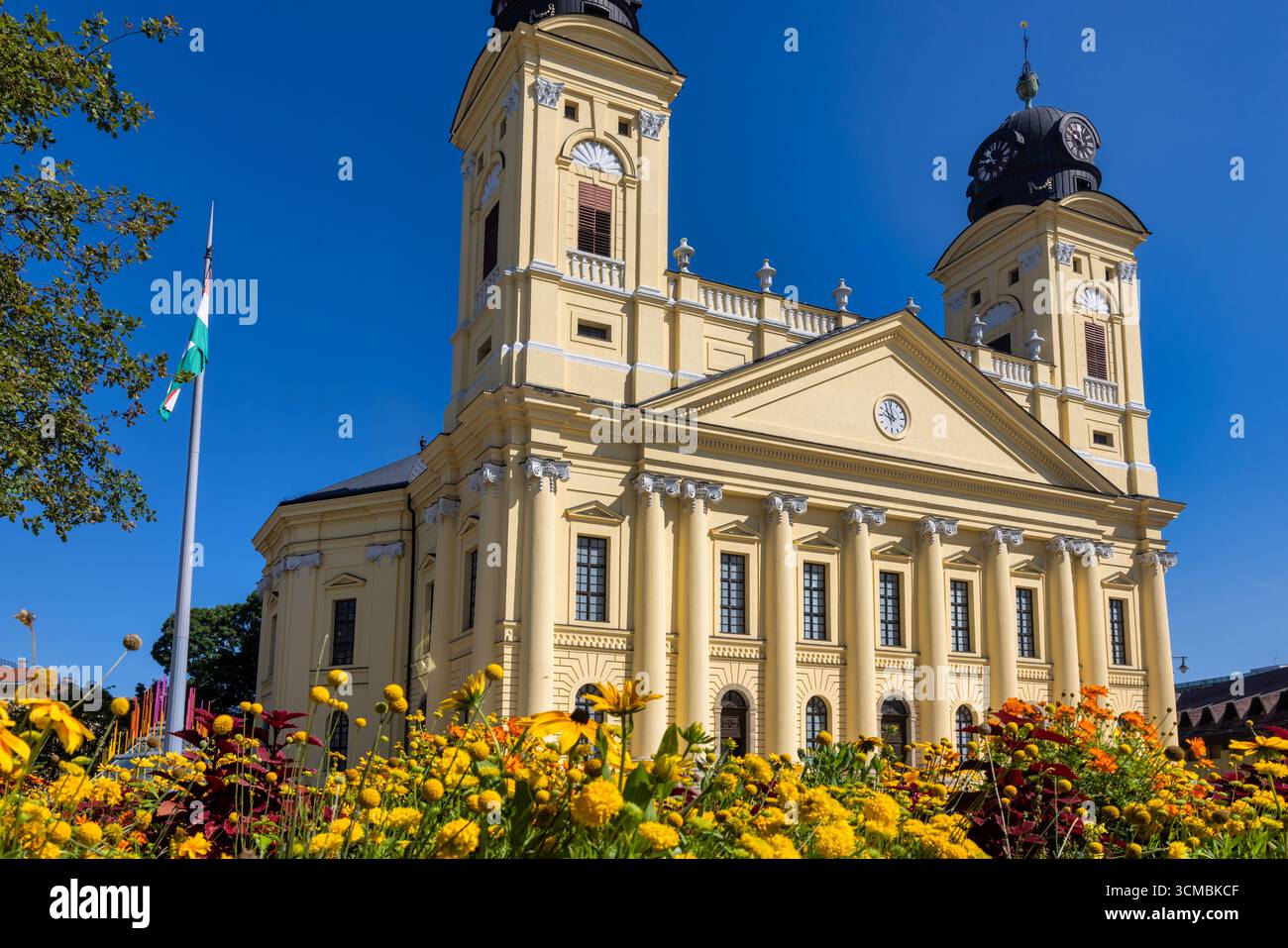 Debrecen, Ungheria 30 luglio 2025: Grande Chiesa riformata con torri gemelle e facciata neoclassica. Fiori estivi colorati fioriscono in primo piano con Hung Foto Stock