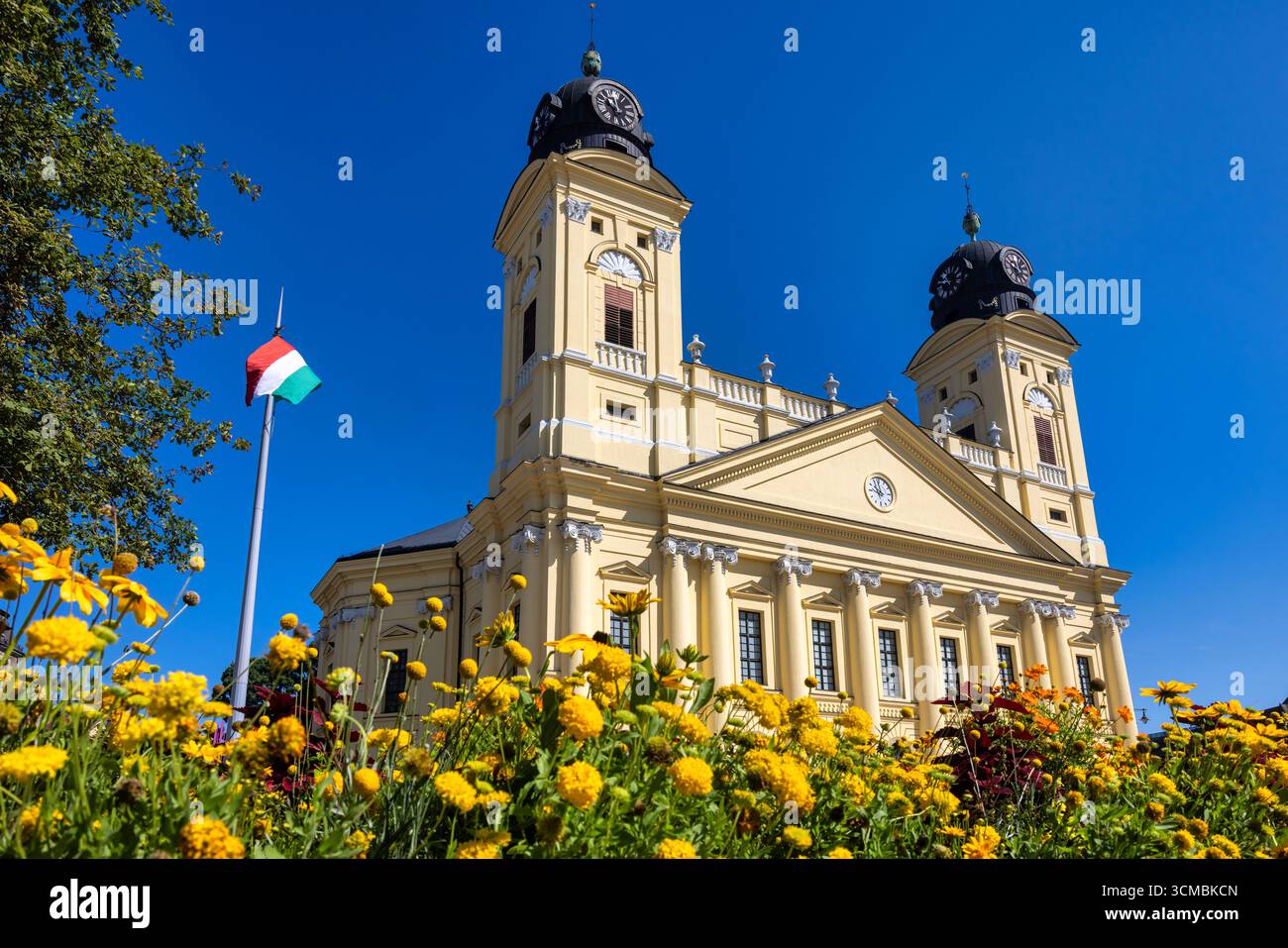 Debrecen, Ungheria 30 luglio 2025: Grande Chiesa riformata con torri gemelle e facciata neoclassica. Fiori estivi colorati fioriscono in primo piano con Hung Foto Stock