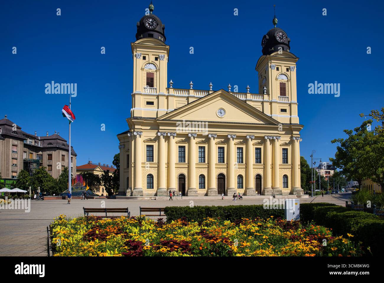 Debrecen, Ungheria 30 luglio 2025: Grande Chiesa riformata con torri gemelle e facciata neoclassica. Fiori estivi colorati fioriscono in primo piano con Hung Foto Stock
