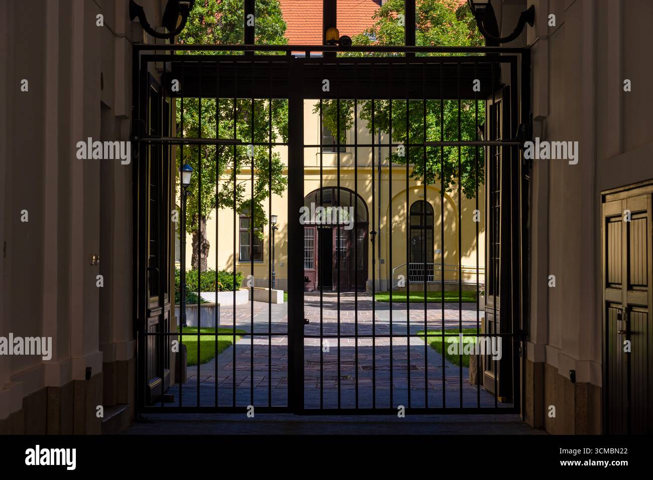 Debrecen, Ungheria 30 luglio 2025 – Vista attraverso il cancello nero sul cortile illuminato dal sole con finestre ad arco, tetto rosso, alberi e pareti gialle che creano morbide tonalità Foto Stock