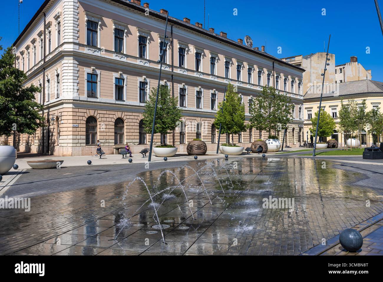 Debrecen, Ungheria 30 luglio 2025: Moderna piazza con fontane con edifici storici sullo sfondo con torri dell'orologio e statua monumento nel centro della città Foto Stock