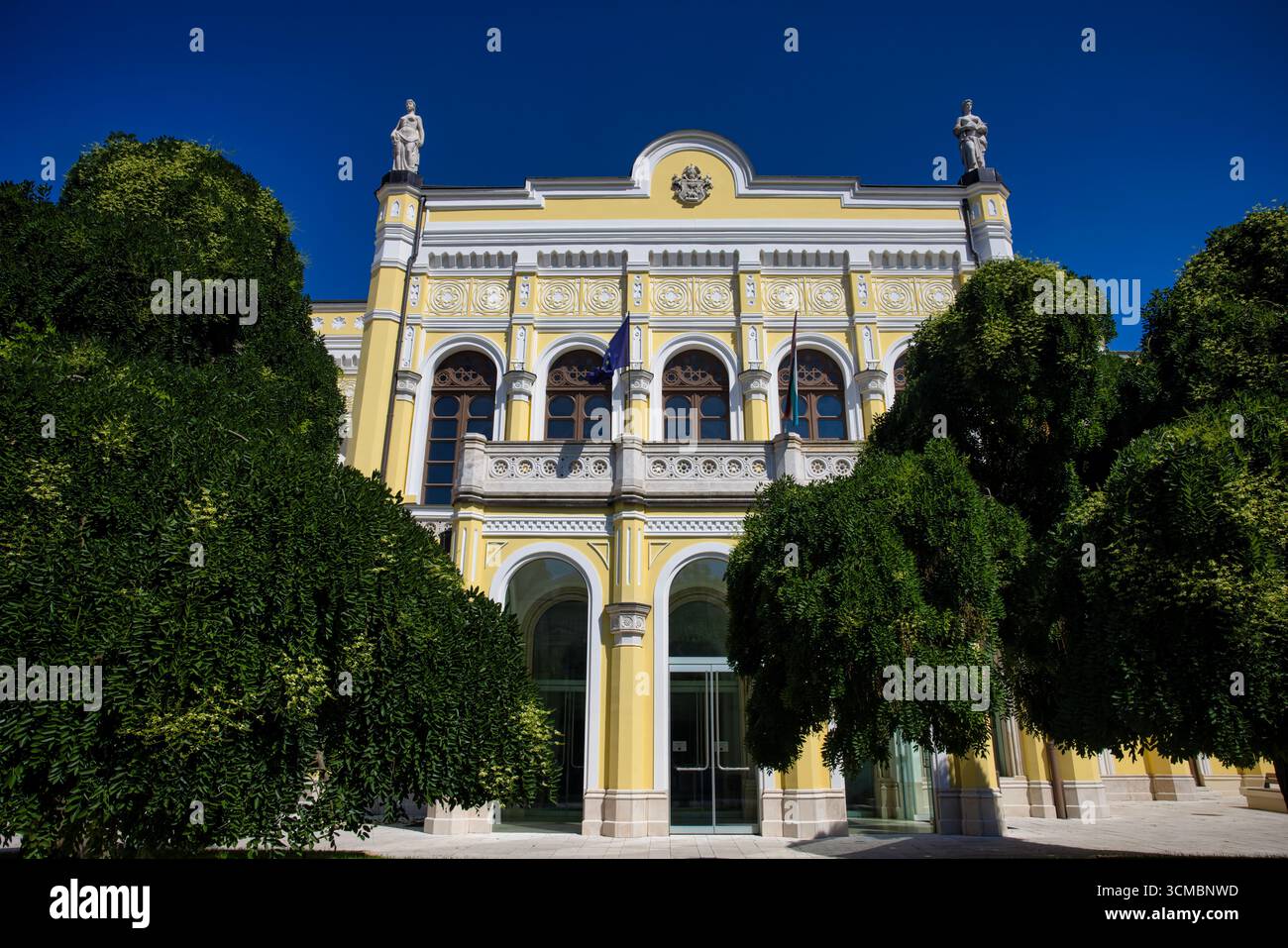 Debrecen, Ungheria 30 luglio 2025 - edificio giallo ornato con statue, cresta, finestre ad arco e colonne, incorniciato da alberi verdi sotto una S blu brillante Foto Stock