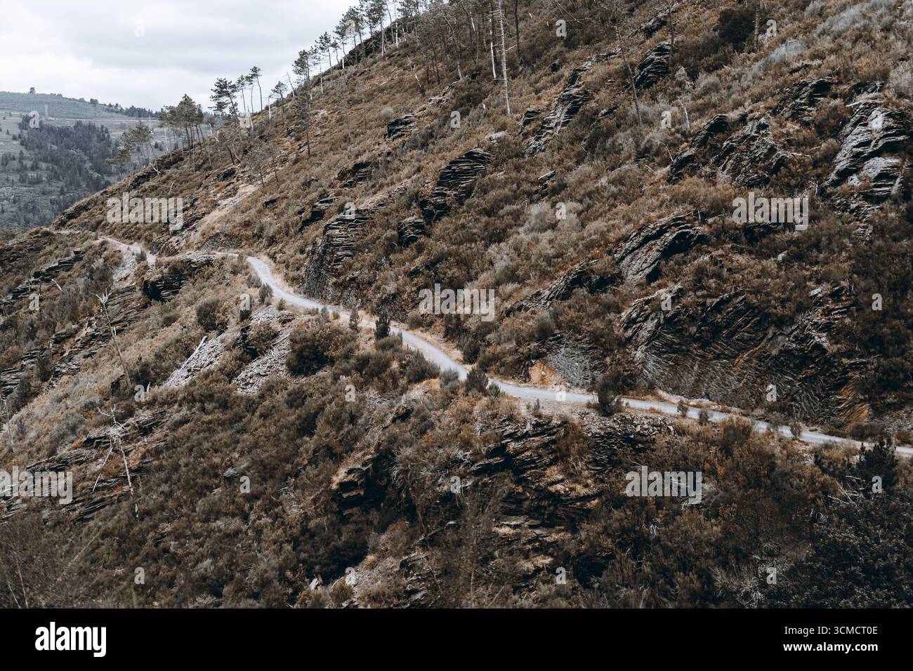 Strada sterrata stretta e tortuosa che attraversa ripidi pendii rocciosi ricoperti da arbusti secchi, alberi sparsi e formazioni rocciose aspre sotto il nuvoloso sk Foto Stock