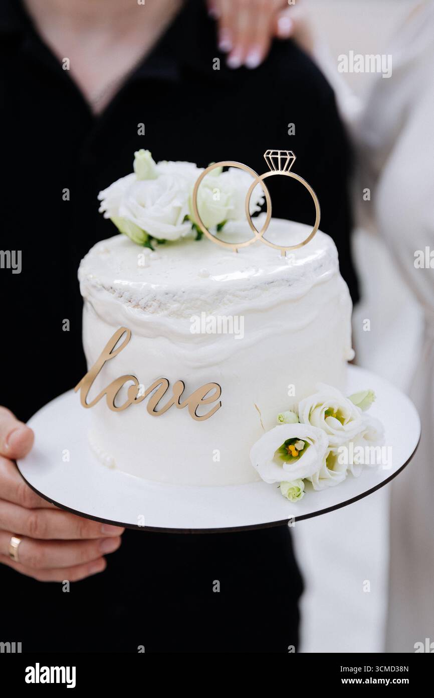 Elegante torta nuziale con anelli e fiori bianchi, tenuta dagli sposi in occasione della celebrazione Foto Stock