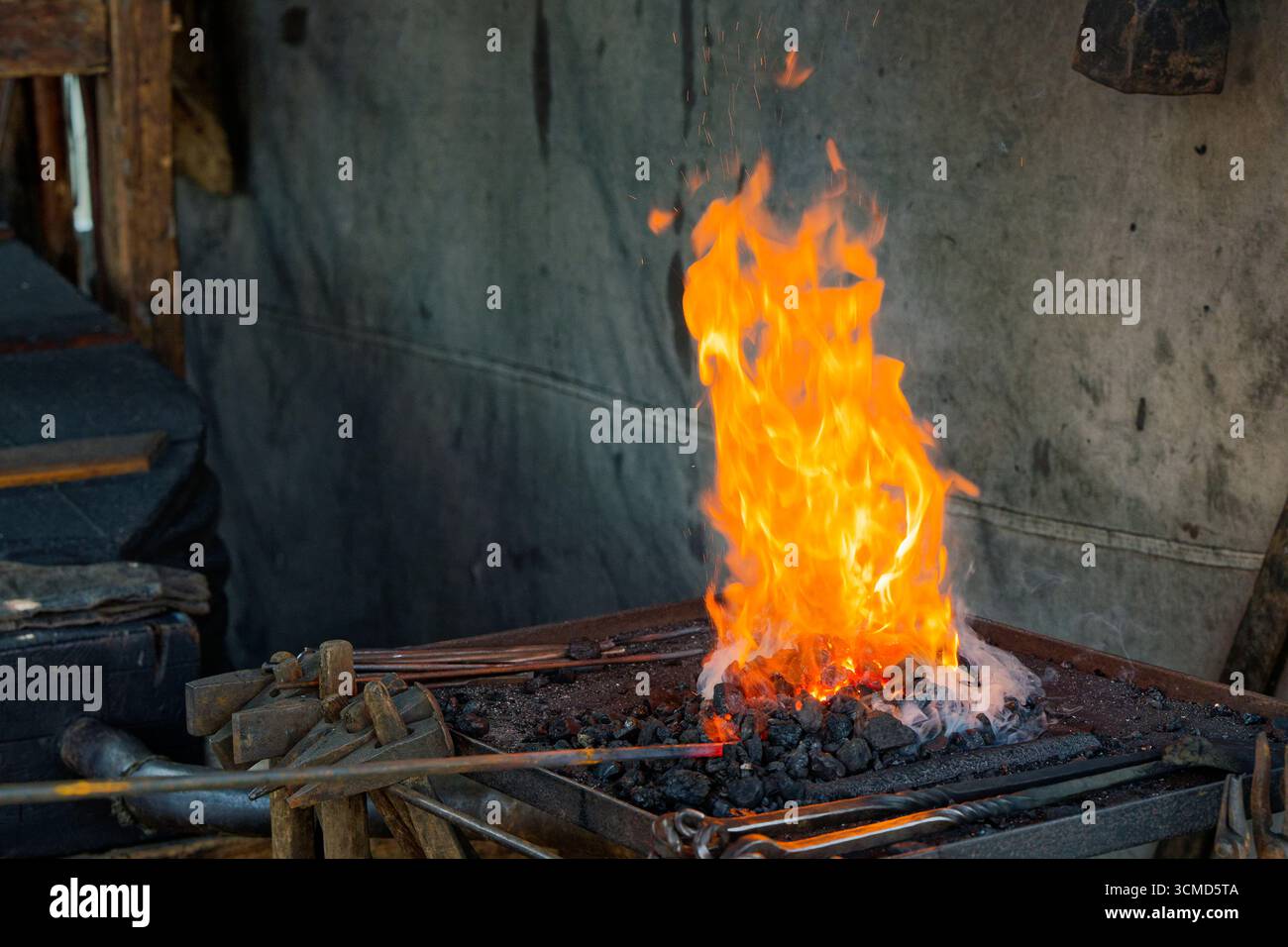 Un focolare metallico pieno di carbone scintillante ruggisce accanto a una parete di cemento. I martelli con manico lungo sono a portata di mano e lo scintillio termico si trova sopra il letto aperto di Foto Stock