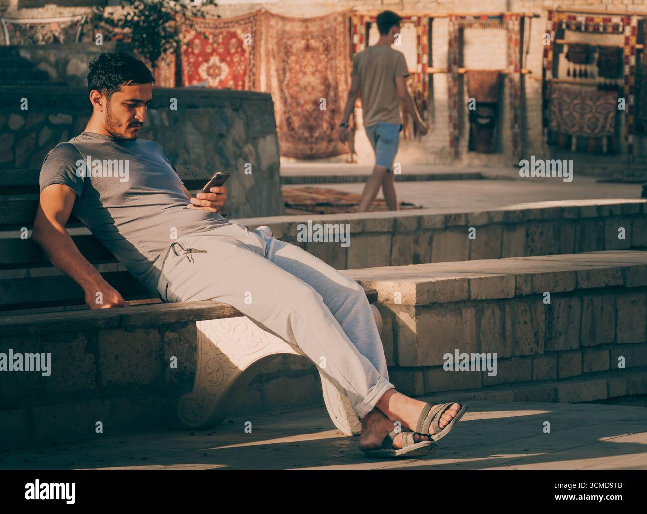 Uomo con camicia grigia e pantaloni bianchi seduto su gradini di pietra, avvolto nel telefono, Bukhara, Uzbekistan, con pareti in mattoni illuminati dal sole e tessuti appesi Foto Stock