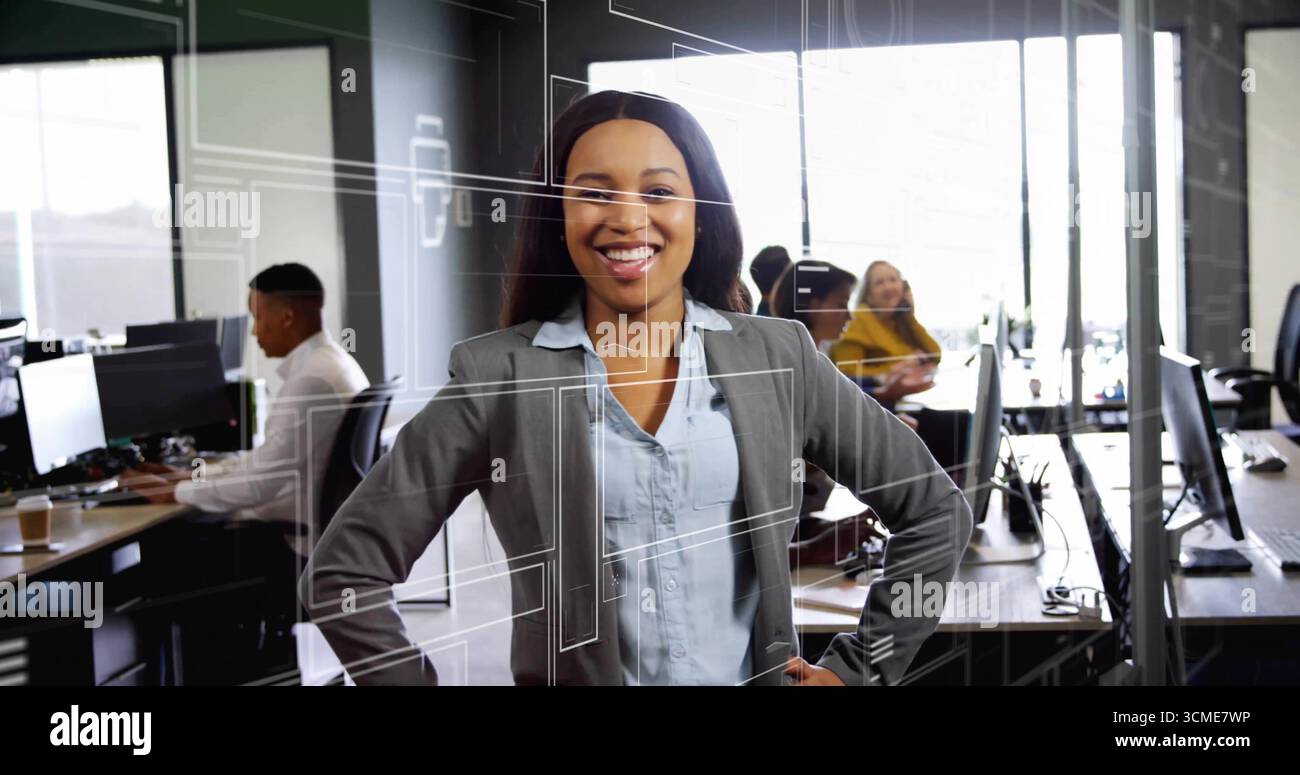 Donna sorridente che indossa un blazer grigio in piedi in ufficio, con scrivanie e computer Foto Stock