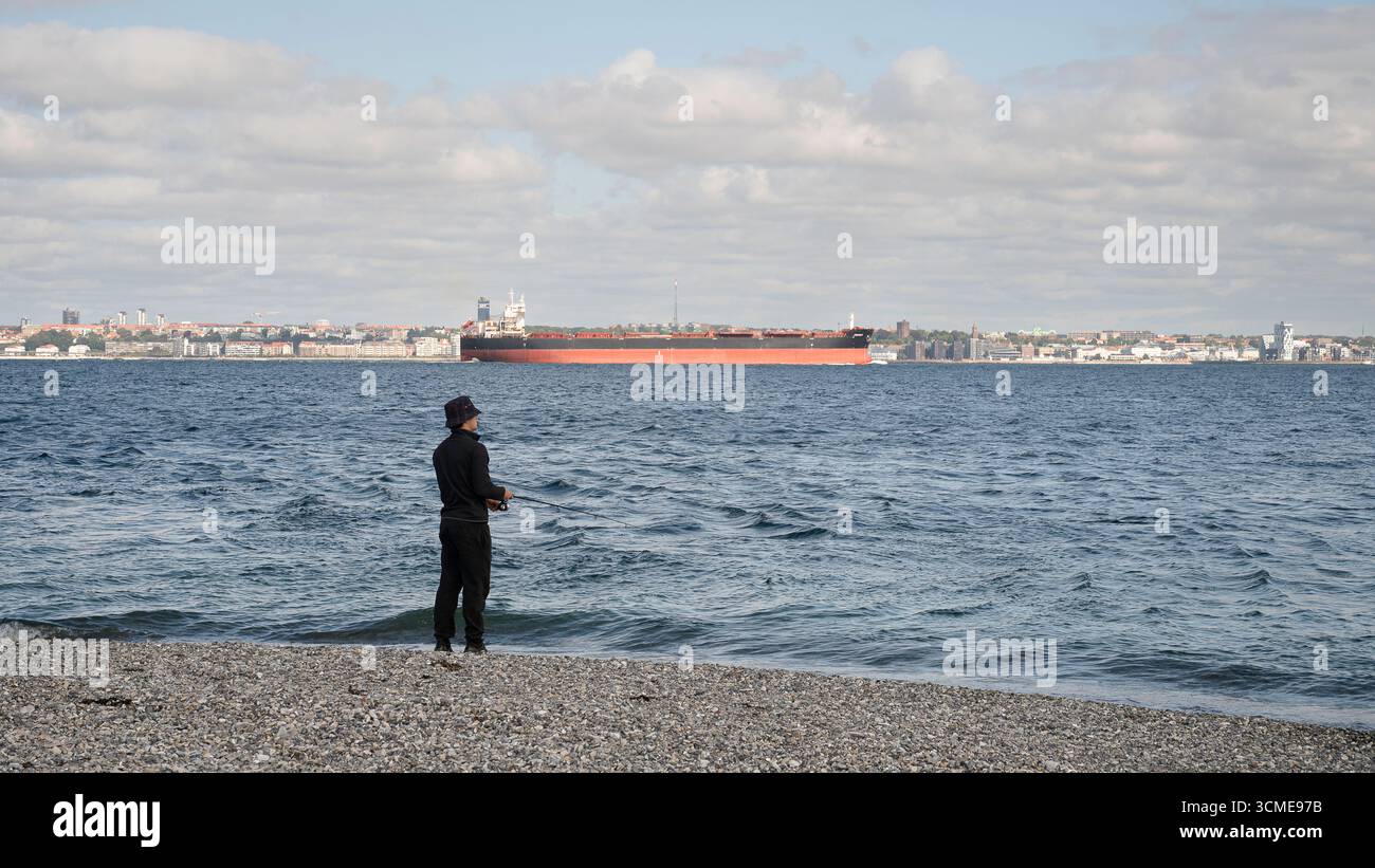 Lo Sound Öresund con un pescatore sulla costa di ciottoli ha trasportato una nave da carico nello stretto di Elsinore, Danimarca, agosto 2025 Foto Stock