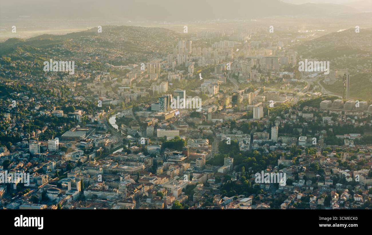 Drone shot dei quartieri moderni di Sarajevo, degli alti edifici e del fiume Miljacka che scorre attraverso la valle sotto la luce nebulosa del tramonto Foto Stock