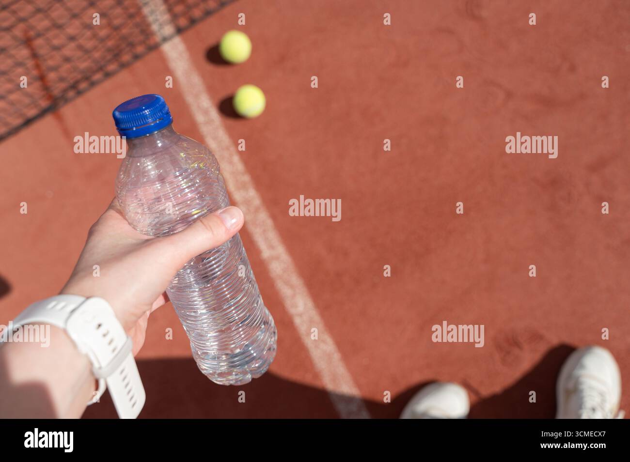 Una persona che tiene abilmente in mano una grande bottiglia d'acqua rinfrescante mentre sta in piedi su un campo da tennis ben tenuto pronto a giocare Foto Stock