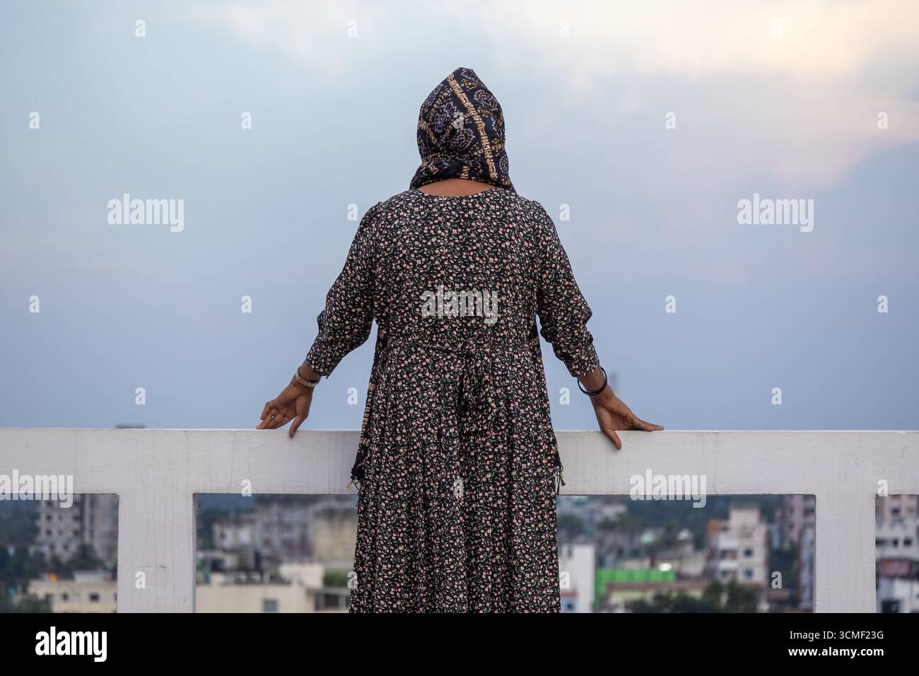 Vista posteriore di una donna musulmana bengalese che indossa un abaya e un hijab floreali, in piedi su un tetto nel pomeriggio, guardando la città e la natura. Foto Stock