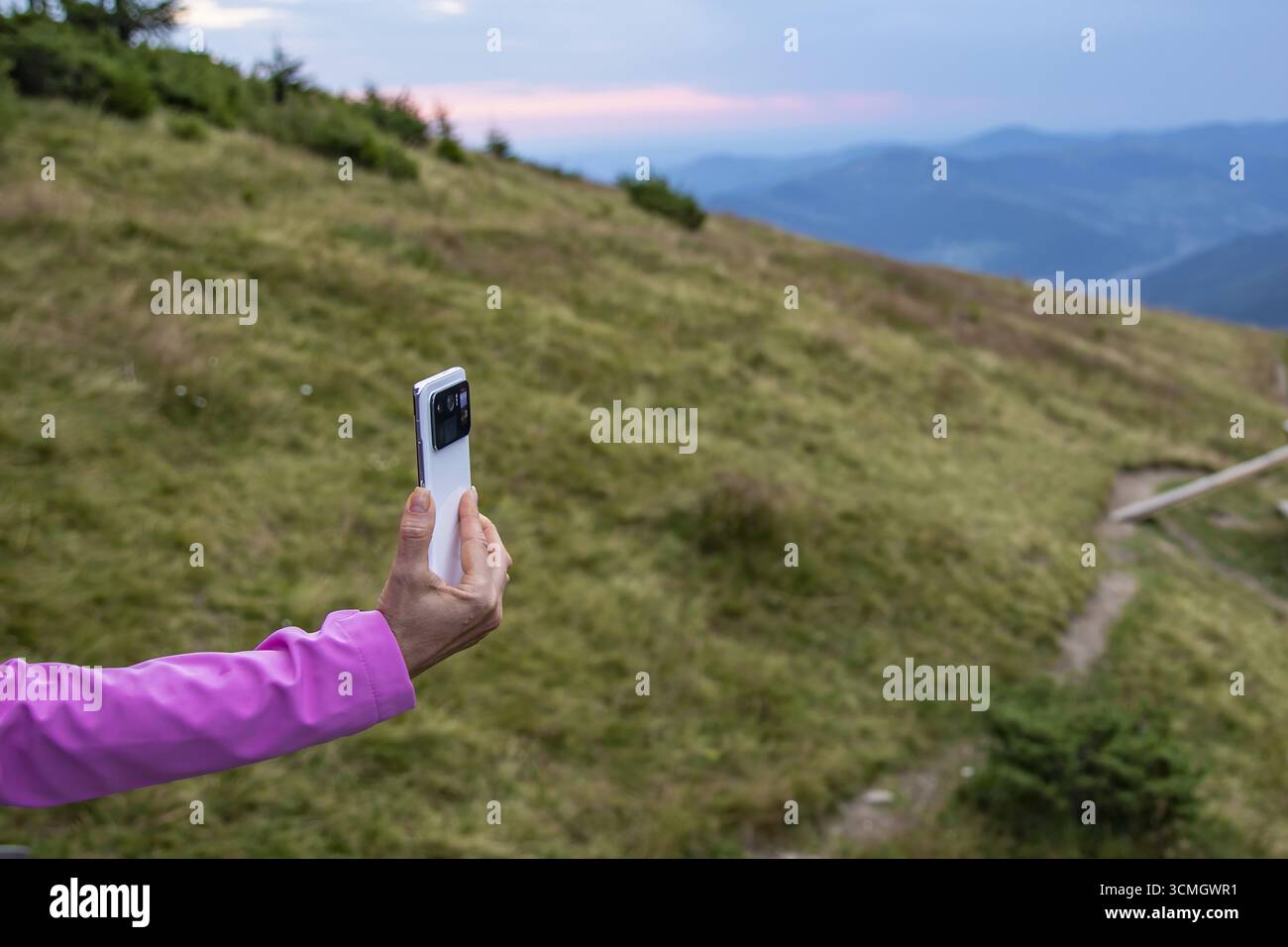 Primo piano di una mano con una giacca rosa che regge lo smartphone sul paesaggio panoramico di montagna dei Carpazi al tramonto. Concetto di viaggio, blogging, digitale Foto Stock