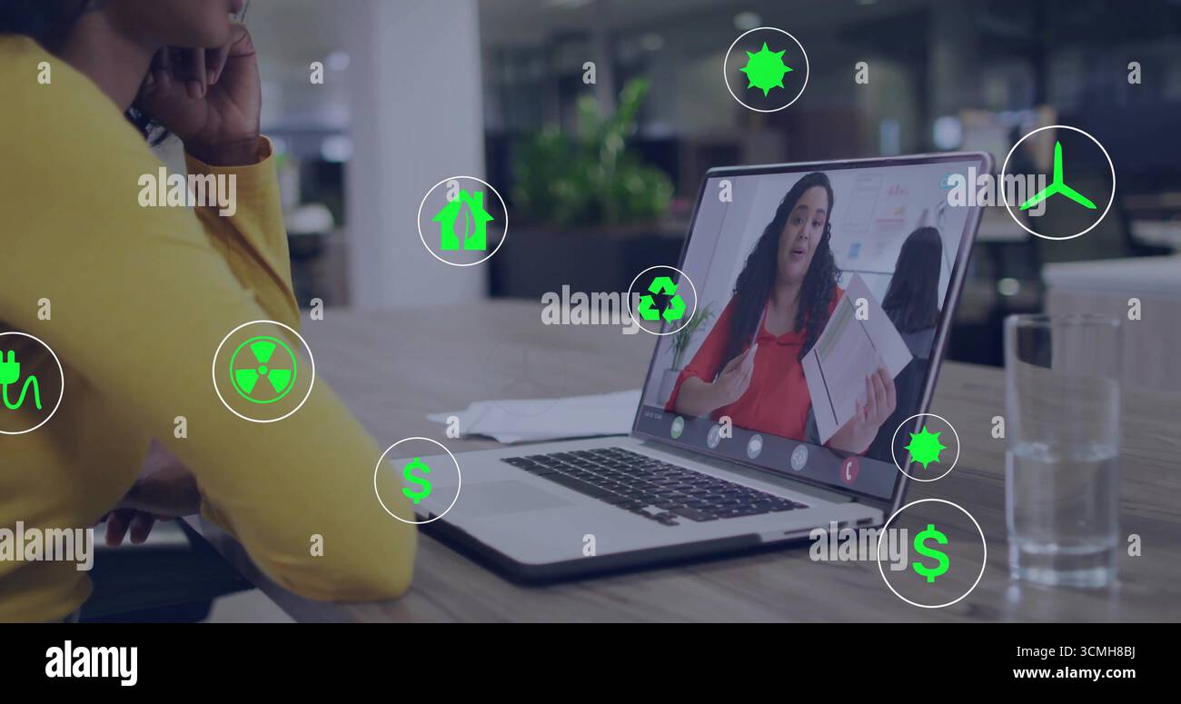 Donna in maglione giallo che guarda una riunione remota in ufficio, con un notebook che mostra icone verdi Foto Stock